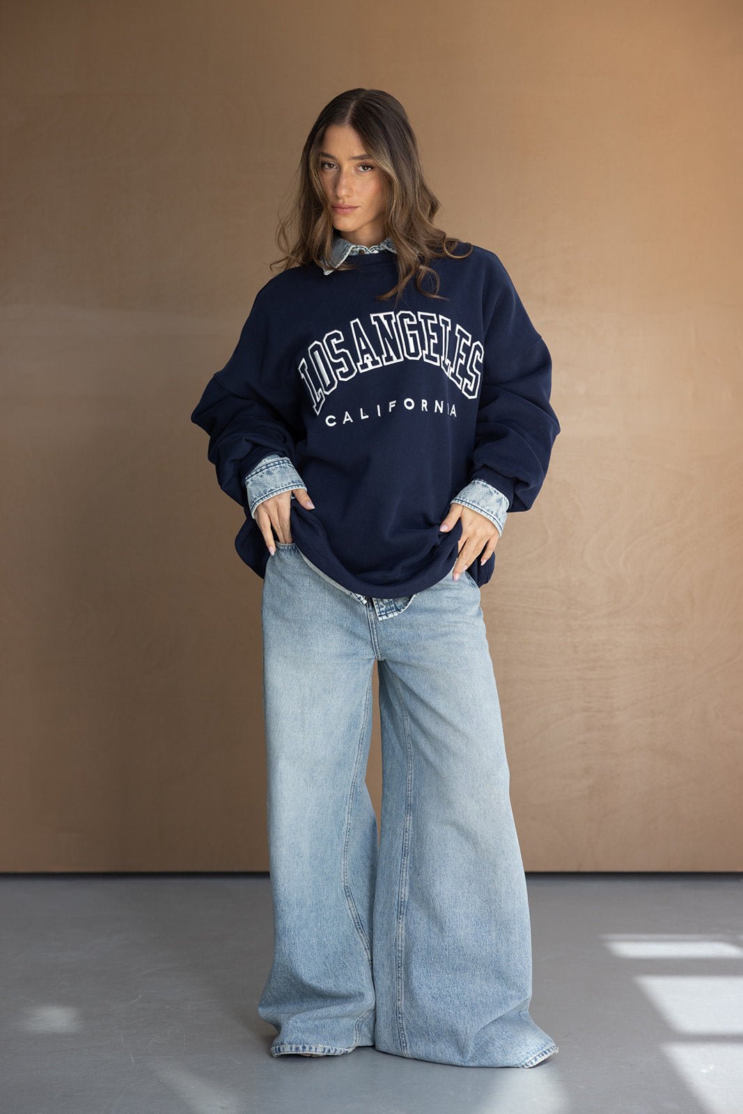 אישה לובשת סווטשירט כחול עם כיתוב 'LOS ANGELES CALIFORNIA' ומכנסי ג'ינס רחבים, אופנת נשים צעירה