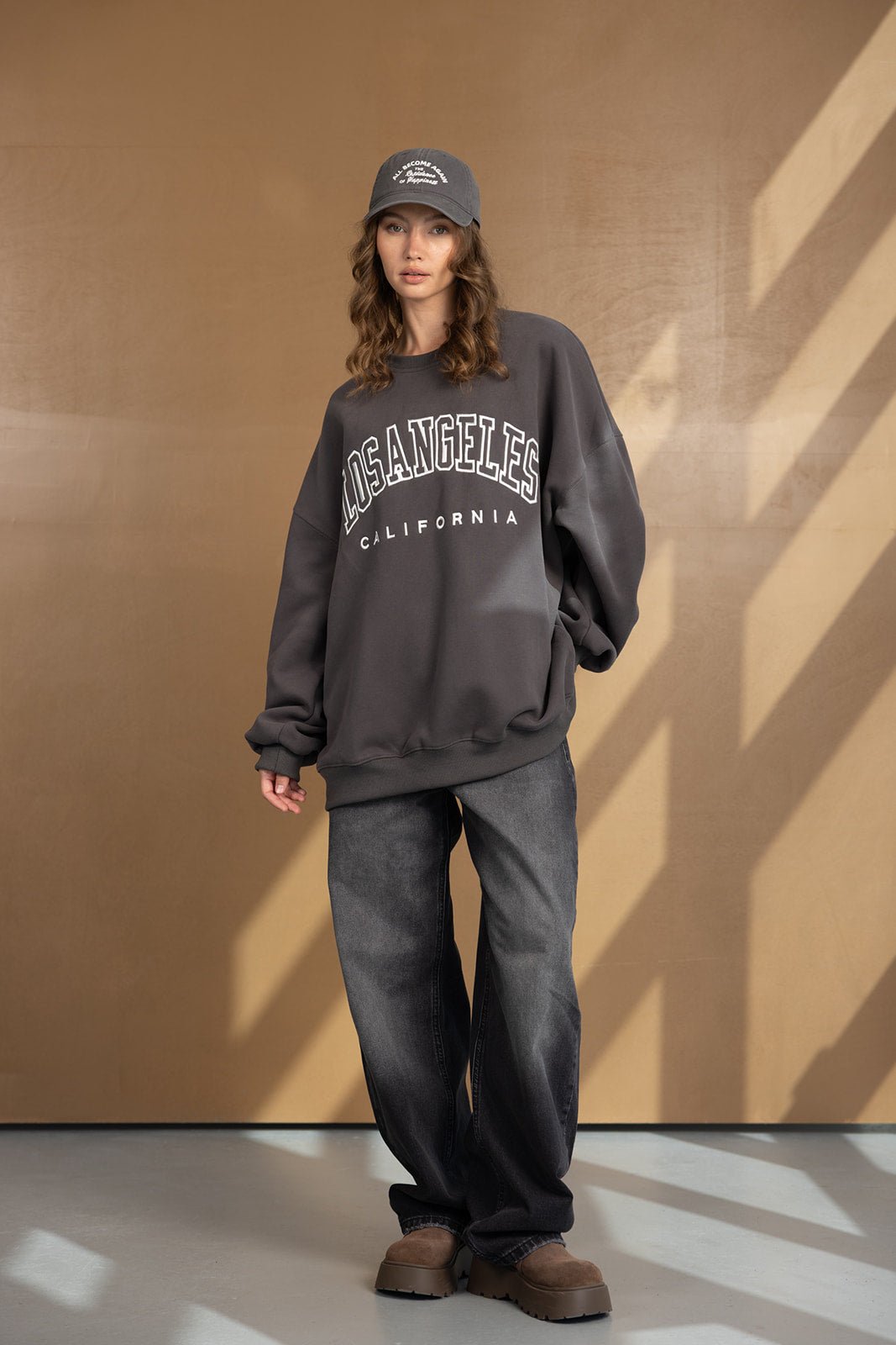 דוגמנית לובשת סווטשירט אפור עם כיתוב 'LOS ANGELES CALIFORNIA', מכנסי ג'ינס רחבים, כובע ונעליים חומות אופנתיות, צילום אופנה לסטייל רחוב לנשים.