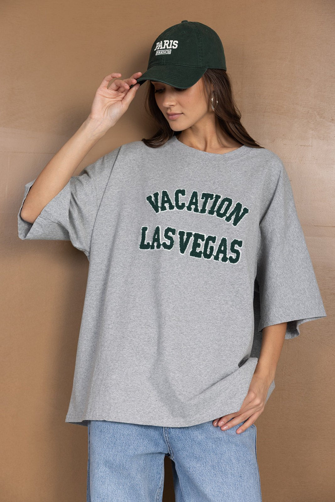 אישה לובשת טישרט אפורה עם כיתוב VACATION LAS VEGAS וכובע ירוק, בגדי קז'ואל לנשים