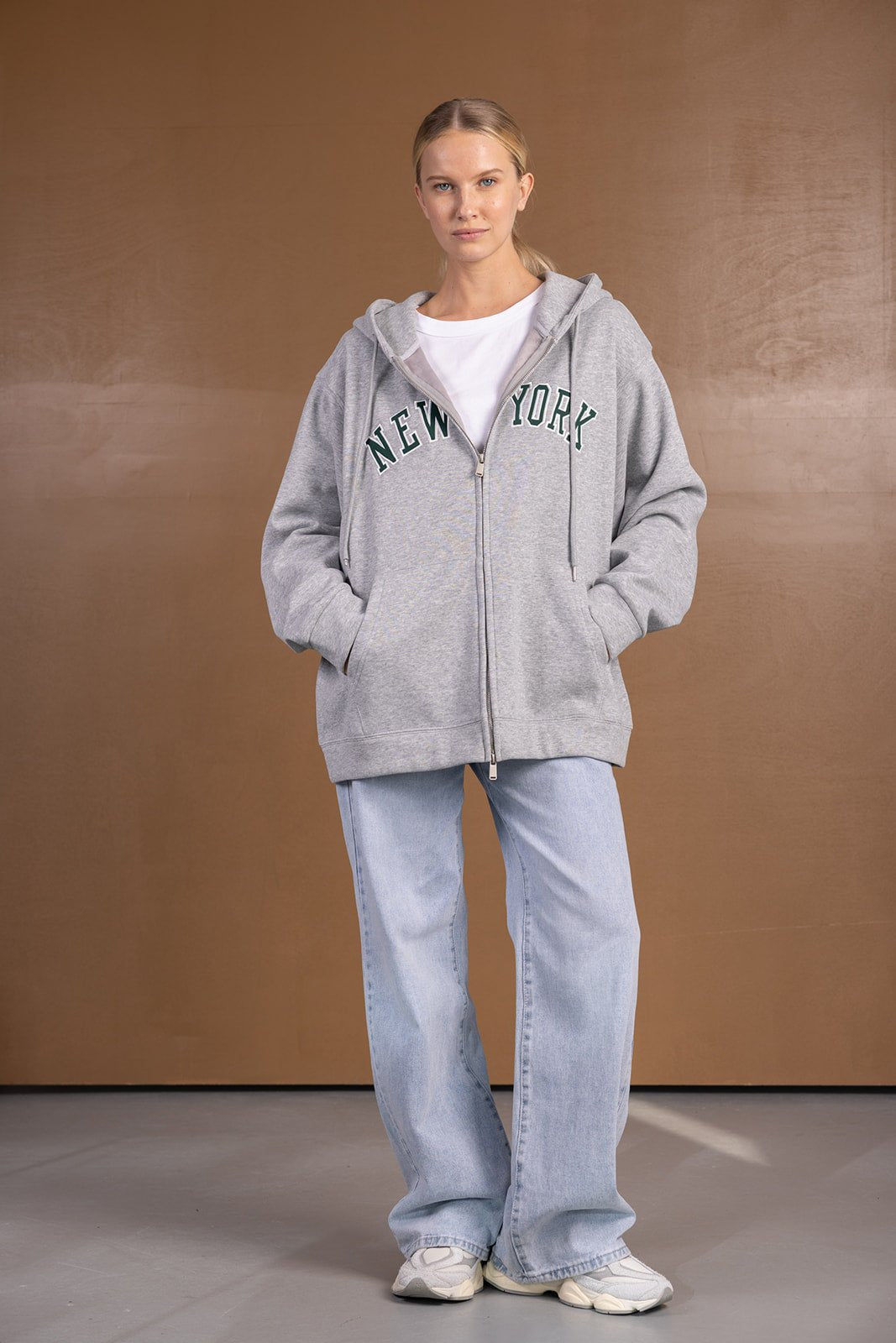 דוגמנית לובשת קפוצ'ון אפור עם כיתוב NEW YORK, ג'ינס בגזרה רחבה ונעלי סניקרס לבנות, אופנת רחוב לנשים
