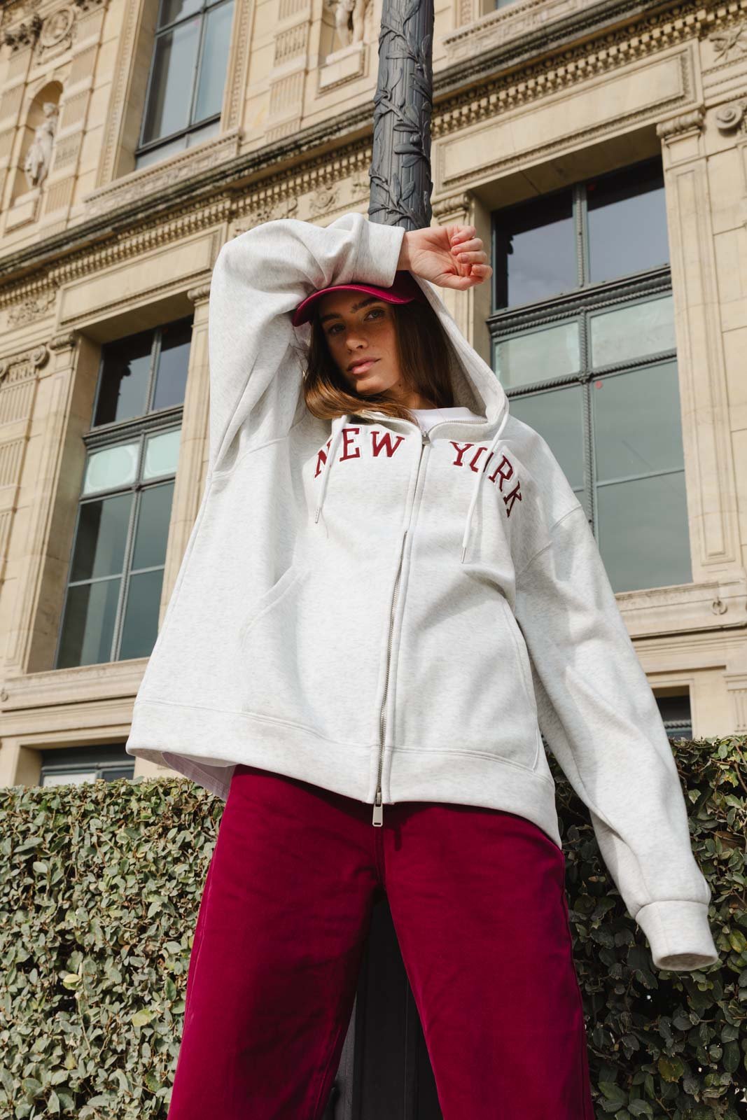 צעירה עם קפוצ׳ון אפור מודפס NEW YORK, כובע מצחייה אדום ומכנסיים בורדו, צילום אופנה אורבני