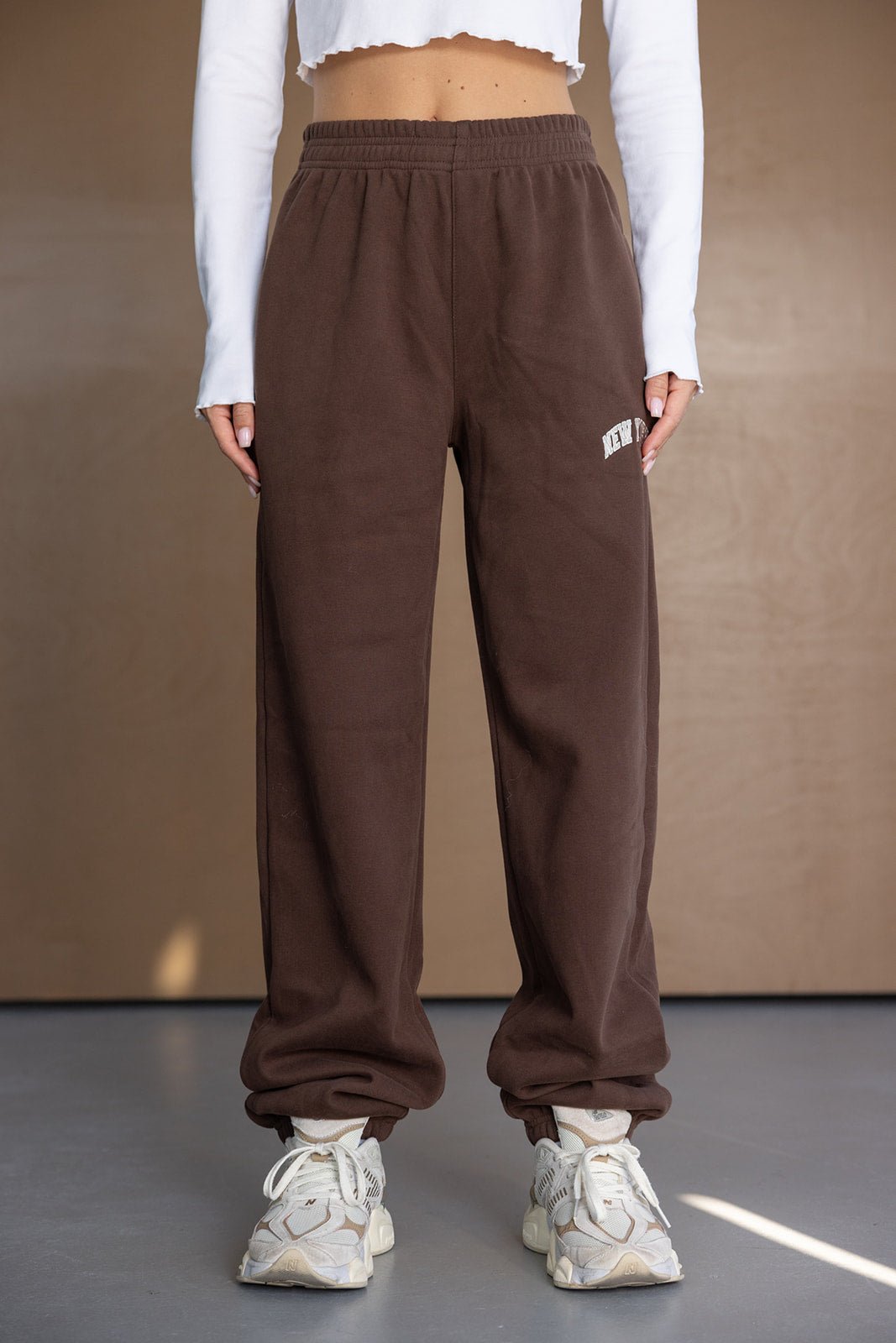 מכנסי טרנינג חומים לנשים עם גומי במותן, חולצת בטן לבנה ונעלי סניקרס אופנתיות