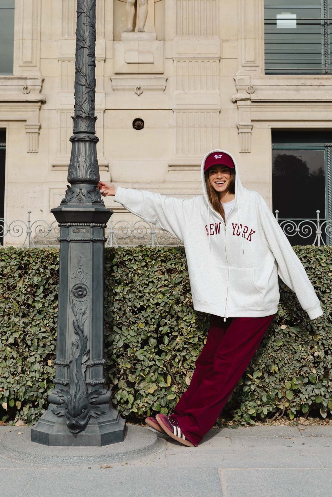 אישה צעירה עם קפוצ'ון לבן וכיתוב NEW YORK, מכנסי טרנינג בורדו וכובע, מצולמת ברחוב עירוני אופנתי
