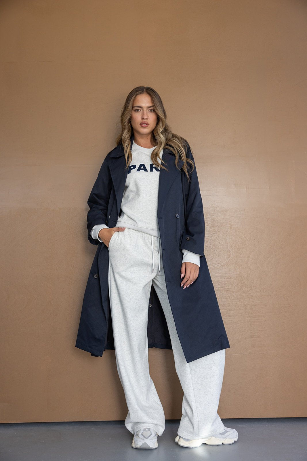 דוגמנית עומדת מול קיר חום עם מעיל טרנץ' כחול כהה, סווטשירט אפור ומכנסיים רחבים בצבע אפרפר, אופנת נשים צעירה וסטייל קז'ואל