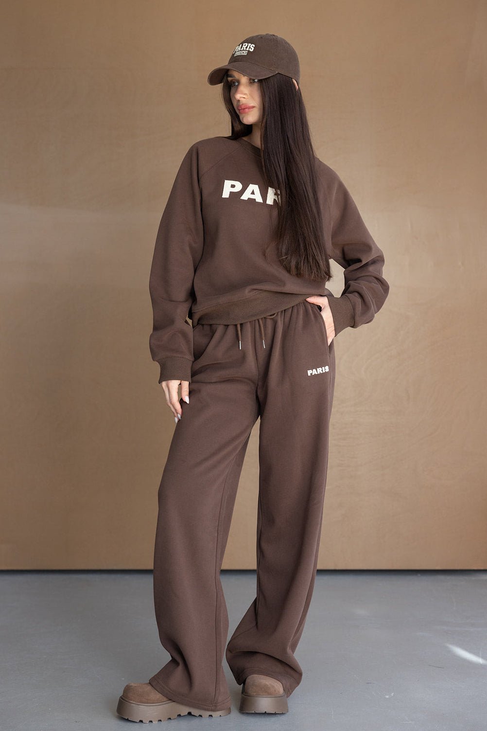 אישה לובשת סט טרנינג חום אופנתי עם כיתוב PARIS, כובע תואם ונעליים שטוחות