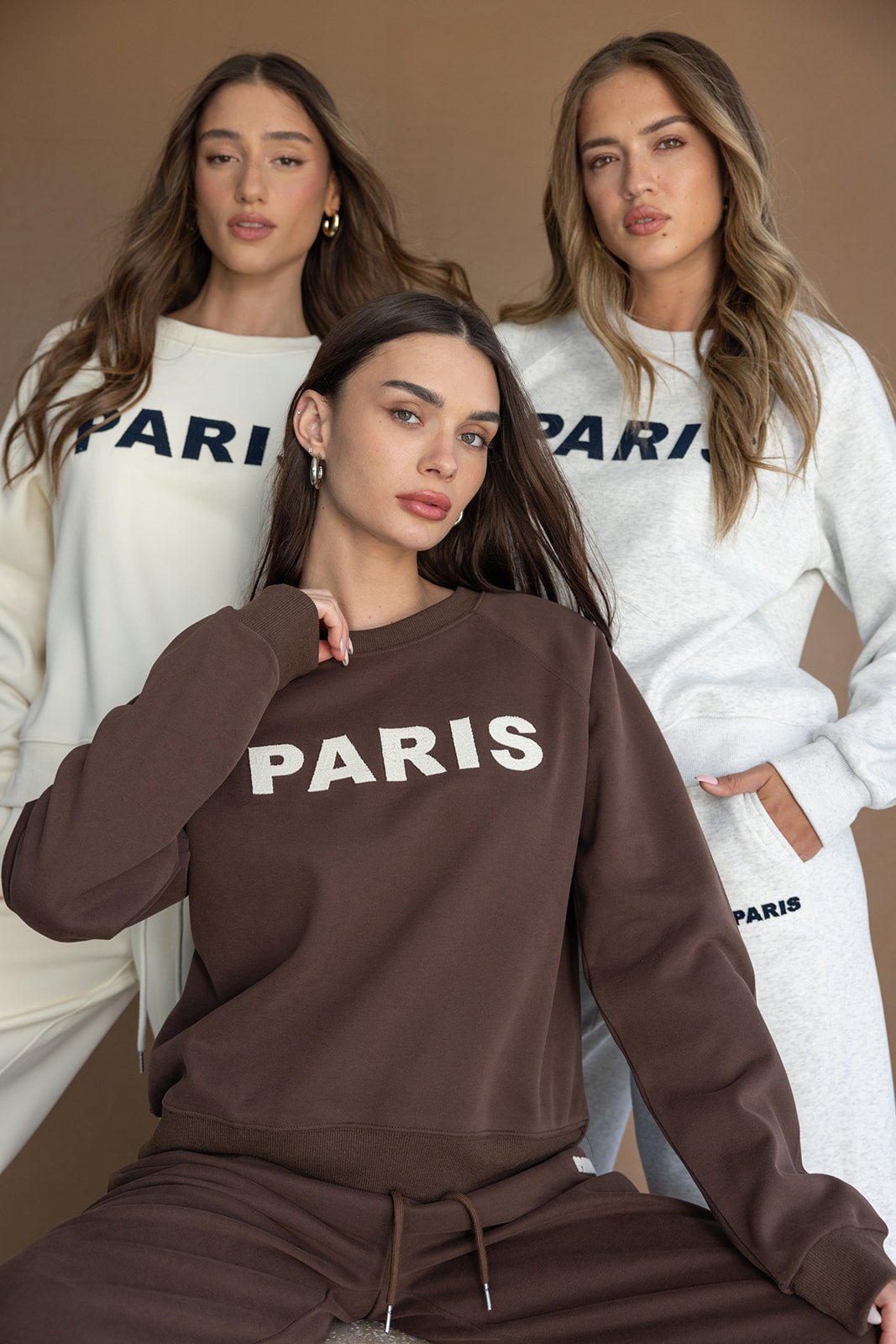 שלוש נשים לובשות חליפות טרנינג אופנתיות עם הדפס PARIS בצבע חום, לבן ואפור