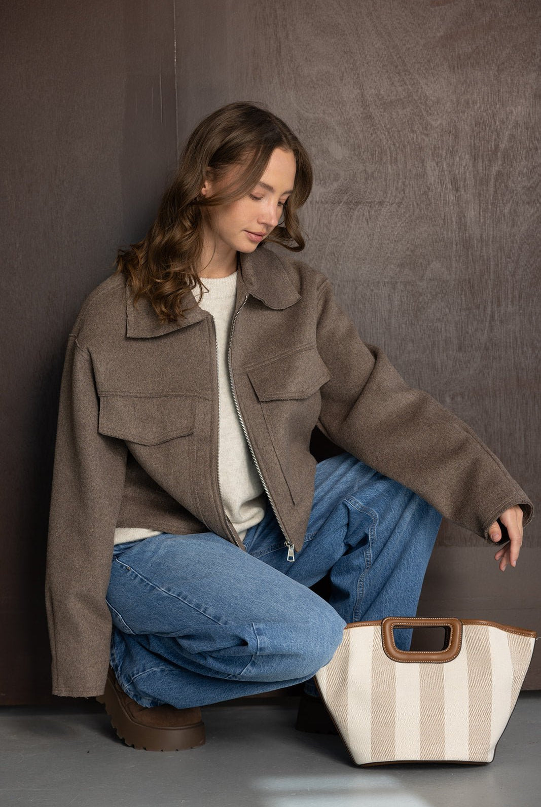 אישה צעירה לובשת ז'קט חום, סוודר בהיר וג'ינס, יושבת עם תיק בד קלאסי, אופנת חורף לנשים