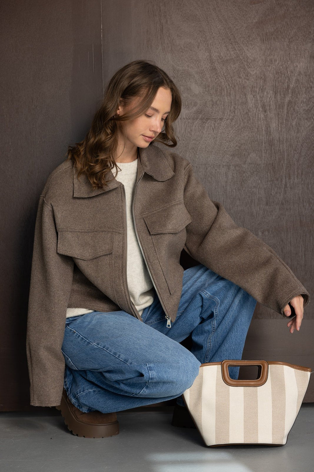 אישה צעירה לובשת ז'קט חום, סוודר בהיר וג'ינס, יושבת עם תיק בד קלאסי, אופנת חורף לנשים