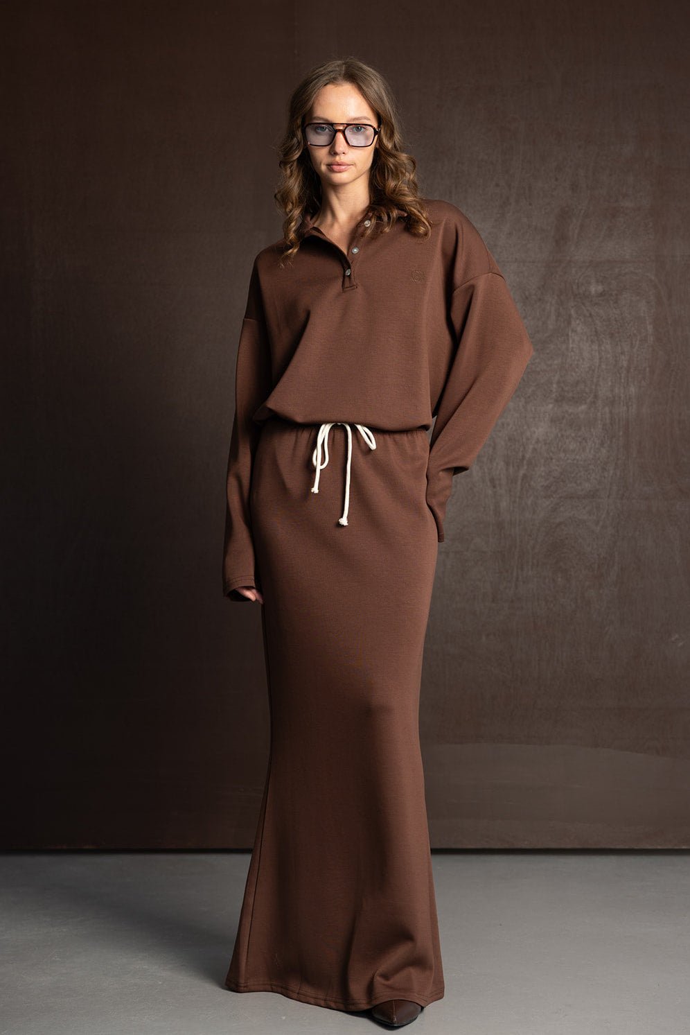 אישה לובשת שמלת מקסי חומה עם שרוך קשירה, משקפיים גדולים, רקע חום, אופנת נשים צעירה