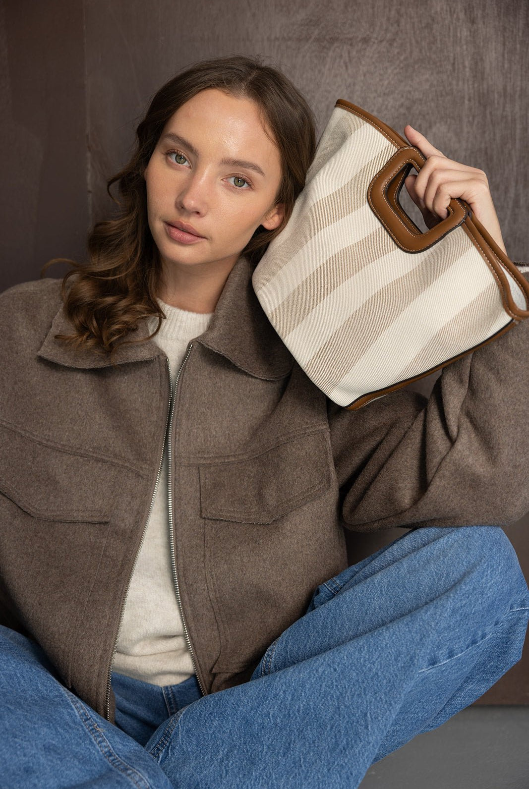 דוגמנית יושבת עם מעיל חום אופנתי, ג'ינס ותיק יד אופנתי בפסים בז' קרם