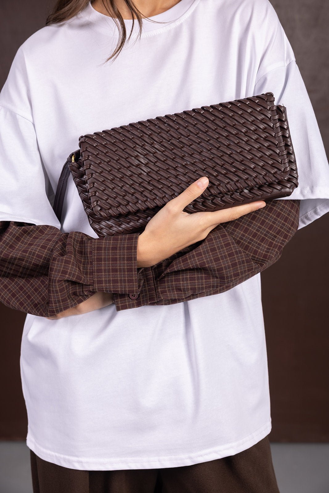 אישה מחזיקה תיק קלאץ' קלוע חום, לובשת חולצה לבנה עם שרוולים משובצים חומים, אופנה לנשים