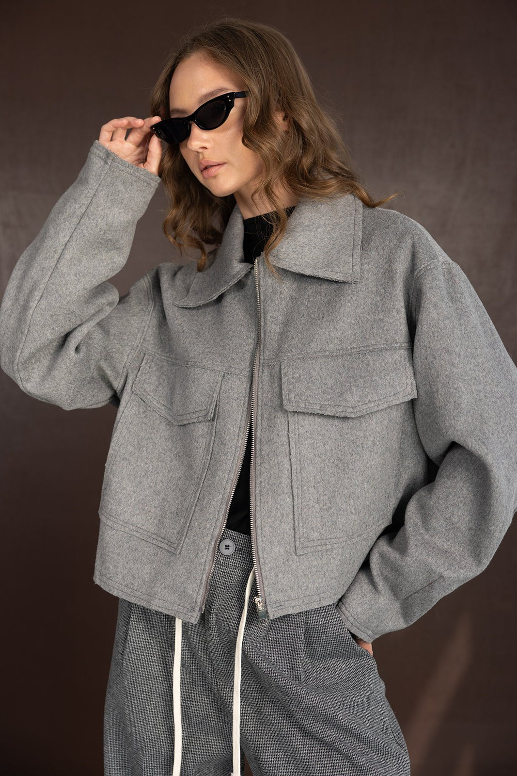 דוגמנית עם מעיל אפור אופנתי, משקפי שמש שחורות ומכנסיים אפורים, אופנת נשים חורפית