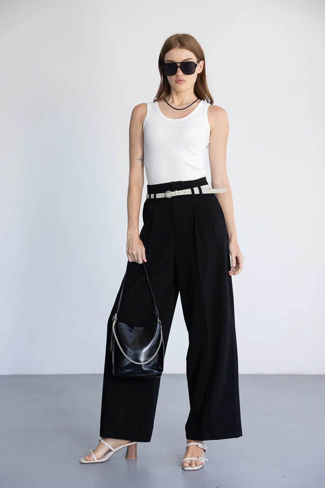 אישה עם גופייה לבנה, מכנסיים שחורים רחבים, חגורה לבנה, תיק שחור ומשקפי שמש, אופנת נשים עכשווית