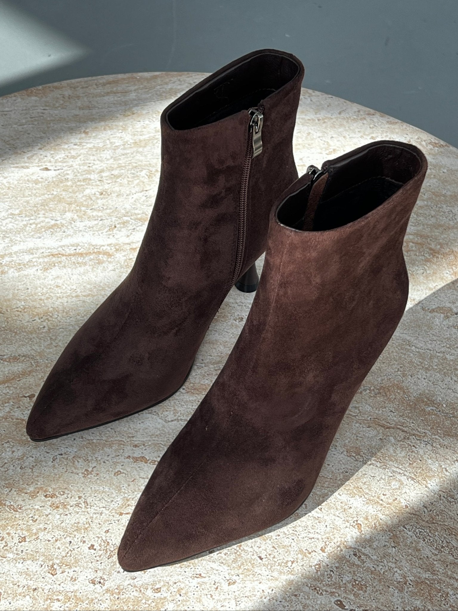 מגפונים חומים לנשים מבד זמש עם עקב גבוה וחוד שפיצי, על שולחן שיש