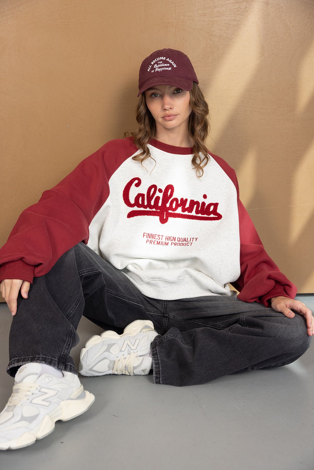 אישה יושבת עם סווטשירט קולור בלוק שרוולים בורדו והדפס California, כובע בייסבול ונעלי סניקרס אופנתיות, לוק קז'ואל צעיר לנשים