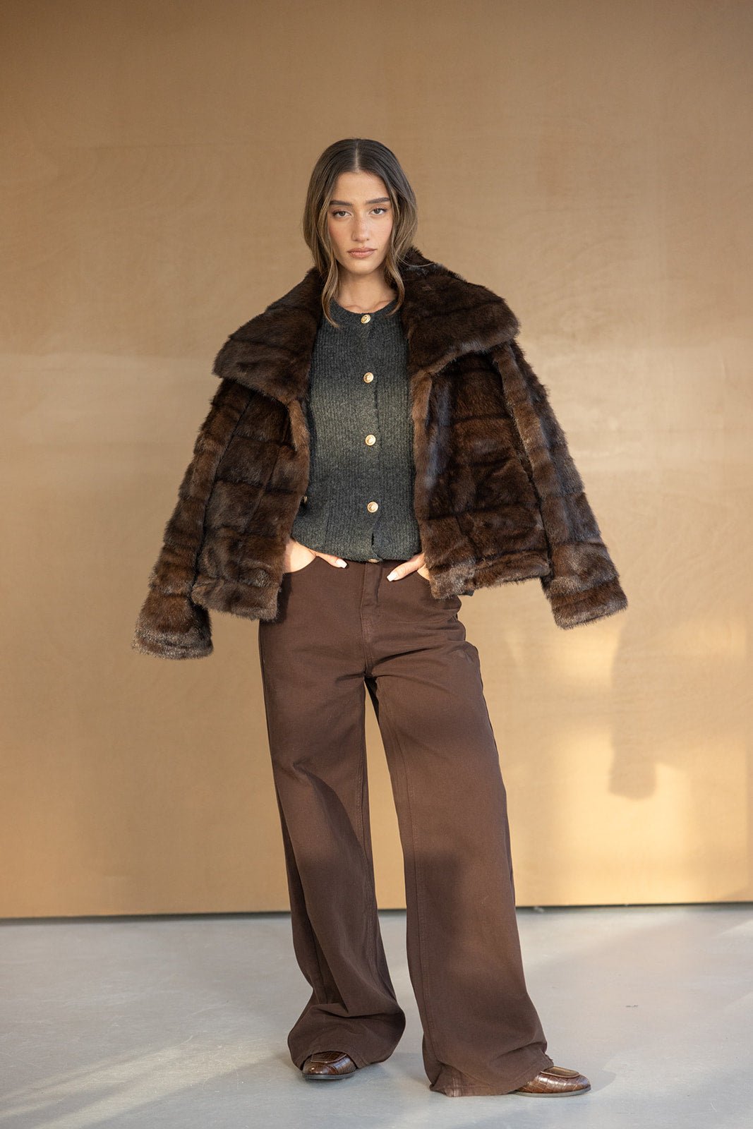 אישה לובשת מעיל פרווה חום, סוודר אפור עם כפתורים ומכנסיים חומים רחבים, אופנת חורף לנשים