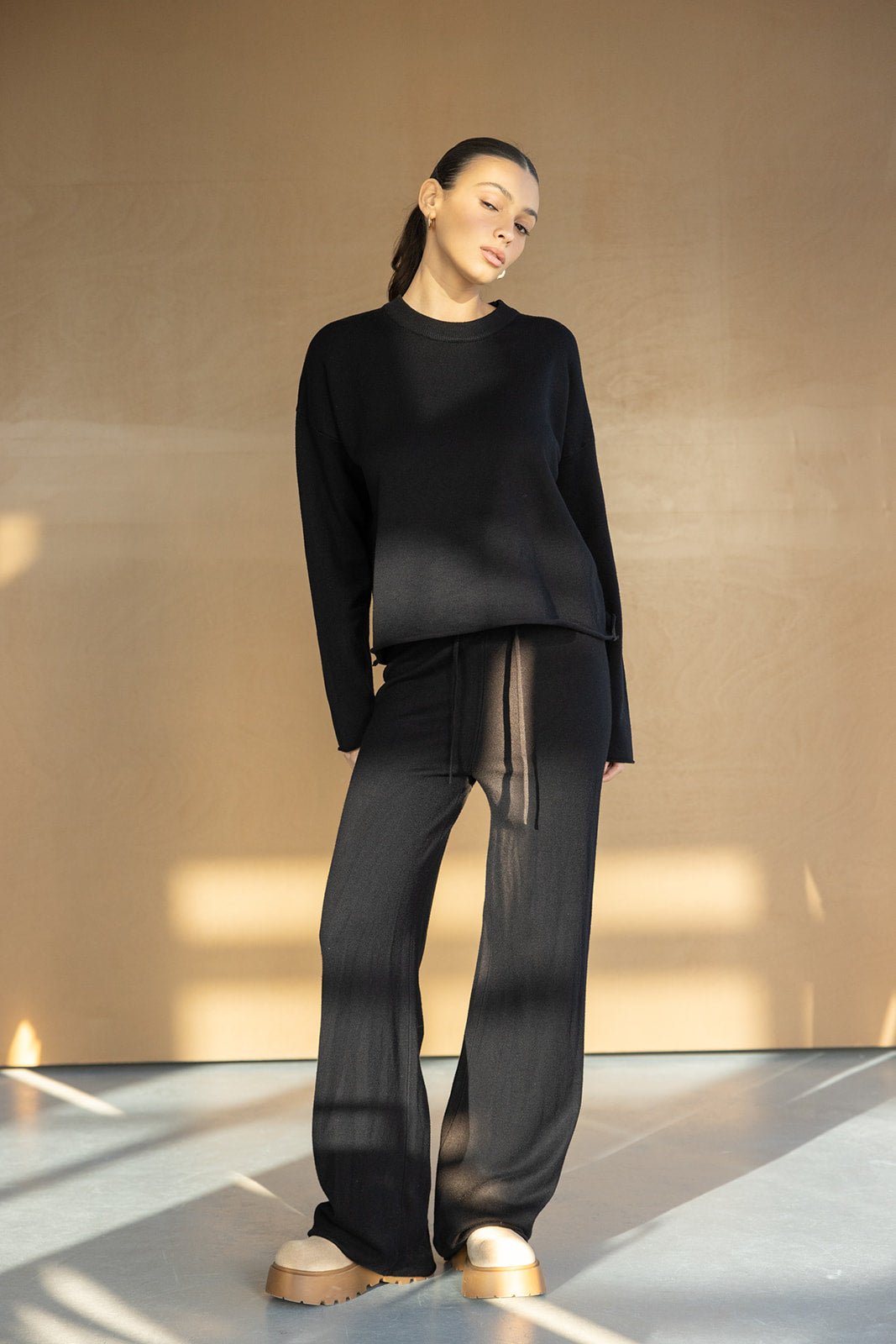 אישה לובשת סט טרנינג שחור אופנתי לנשים, חולצה ארוכה ומכנסיים רחבים, נעלי פלטפורמה בז'.