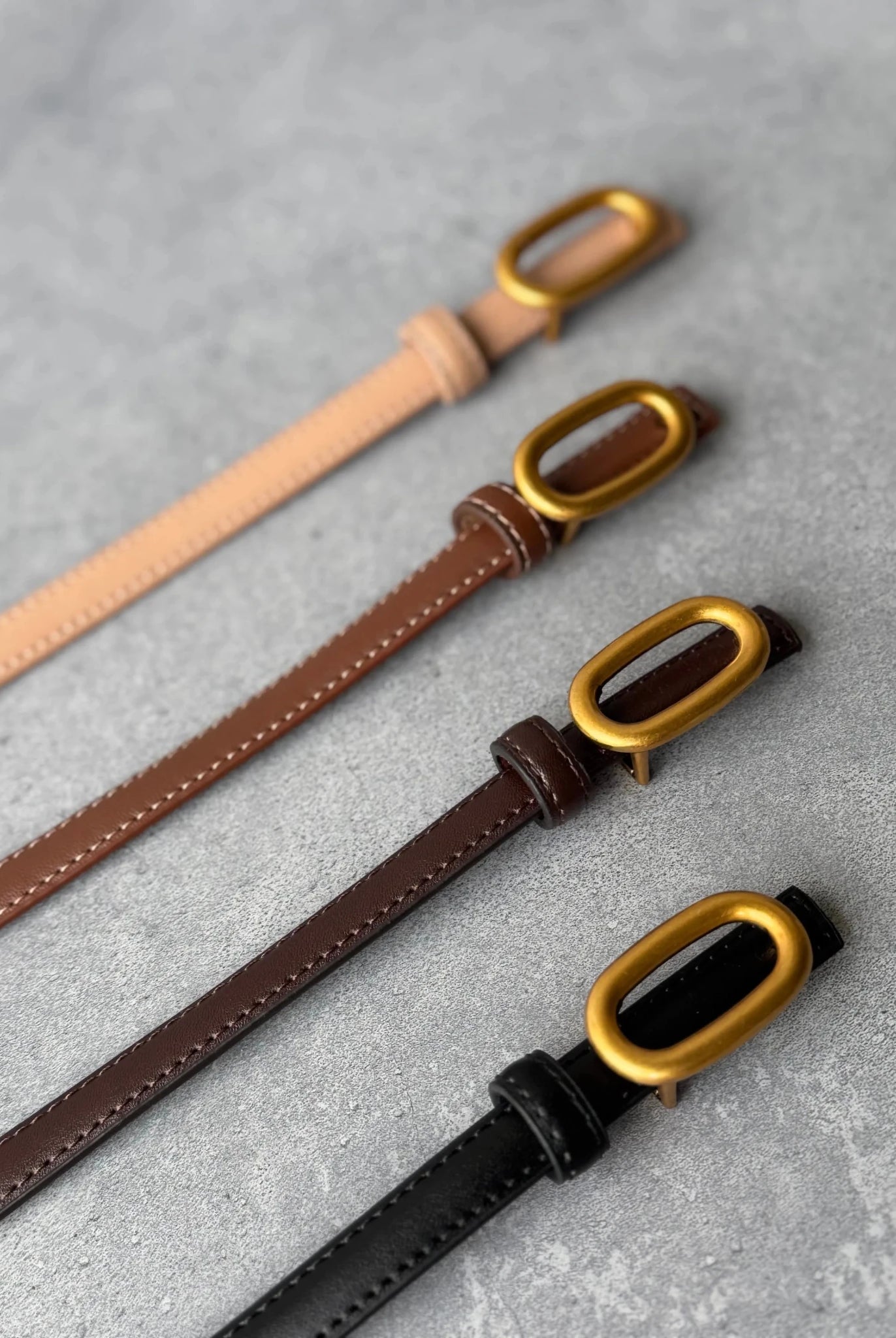 ארבעה חגורות עור בצבע שחור, חום כהה, חום בהיר ובז', אבזם מוזהב, על רקע בטון אפור