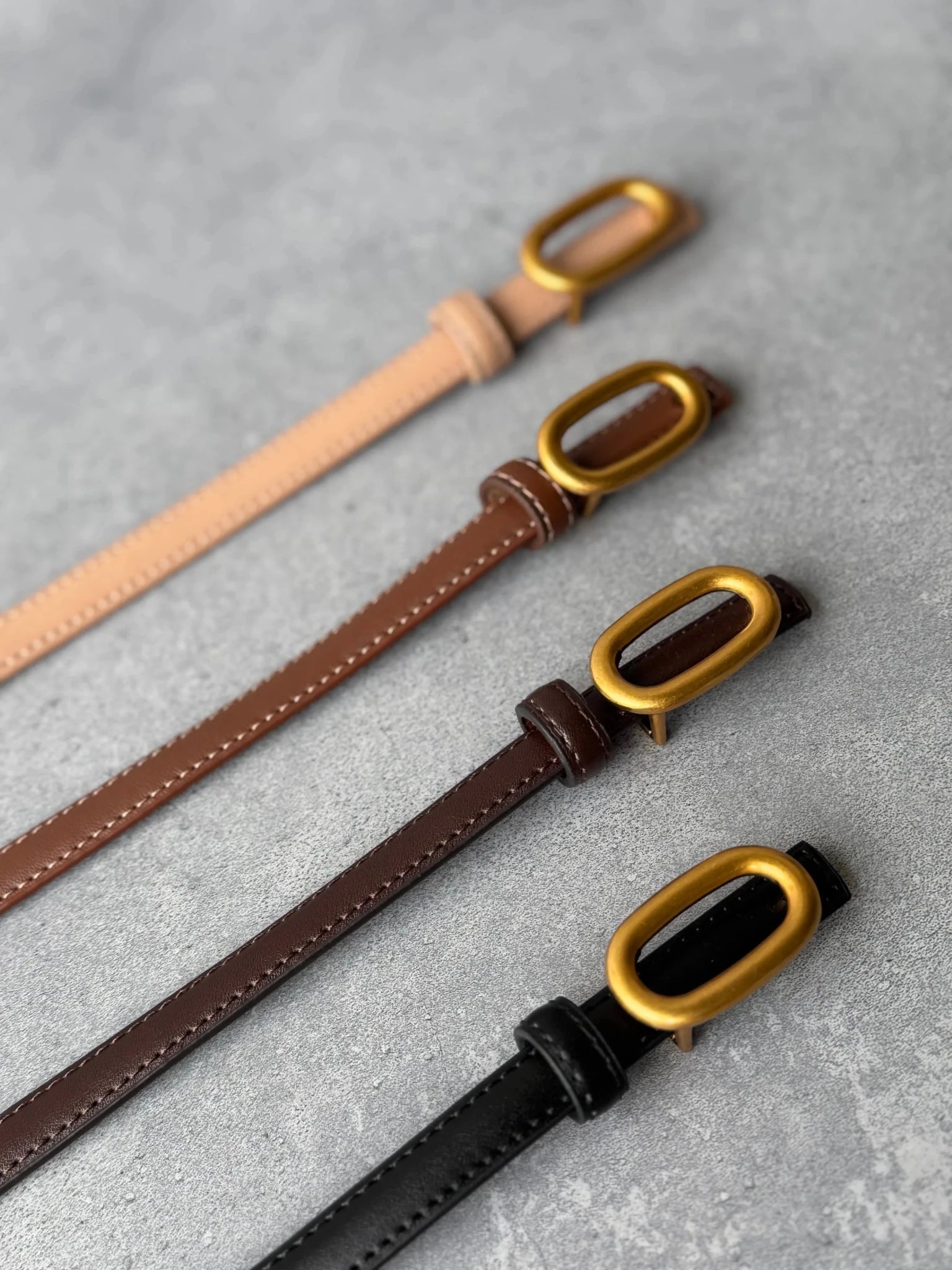 ארבעה חגורות עור בצבע שחור, חום כהה, חום בהיר ובז', אבזם מוזהב, על רקע בטון אפור