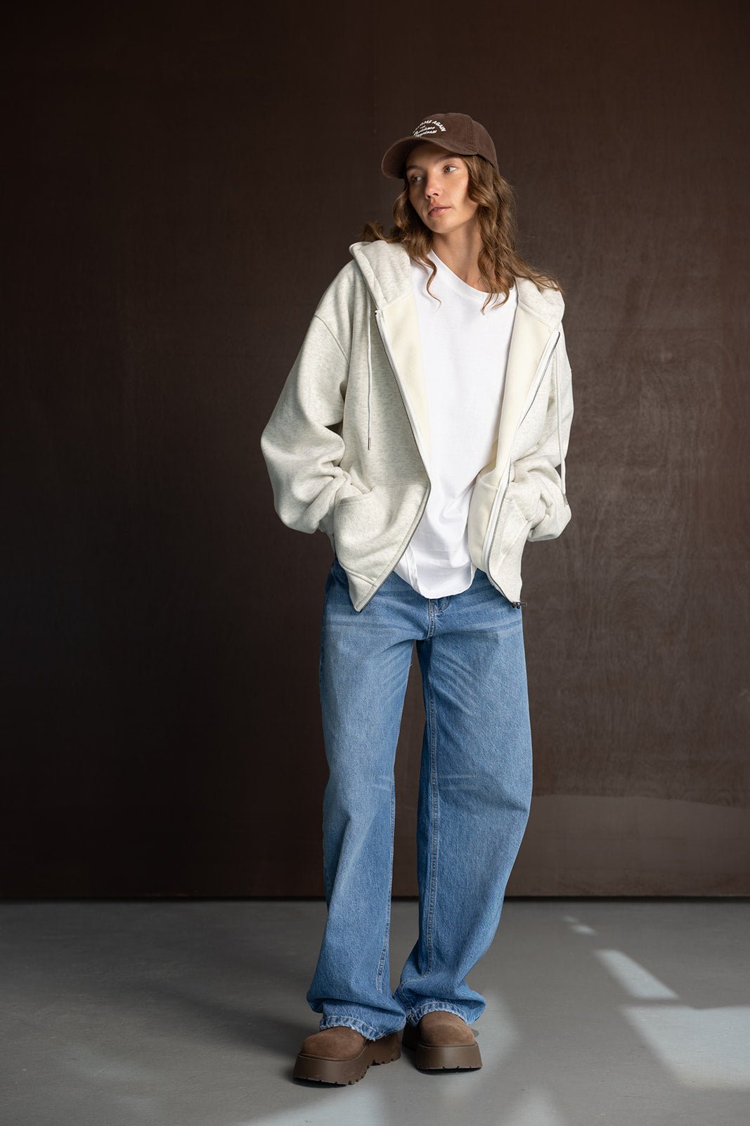 אישה צעירה עם קפוצ'ון אפור, חולצה לבנה, מכנסי ג'ינס רחבים וכובע חום אופנתי לסטייל יומיומי