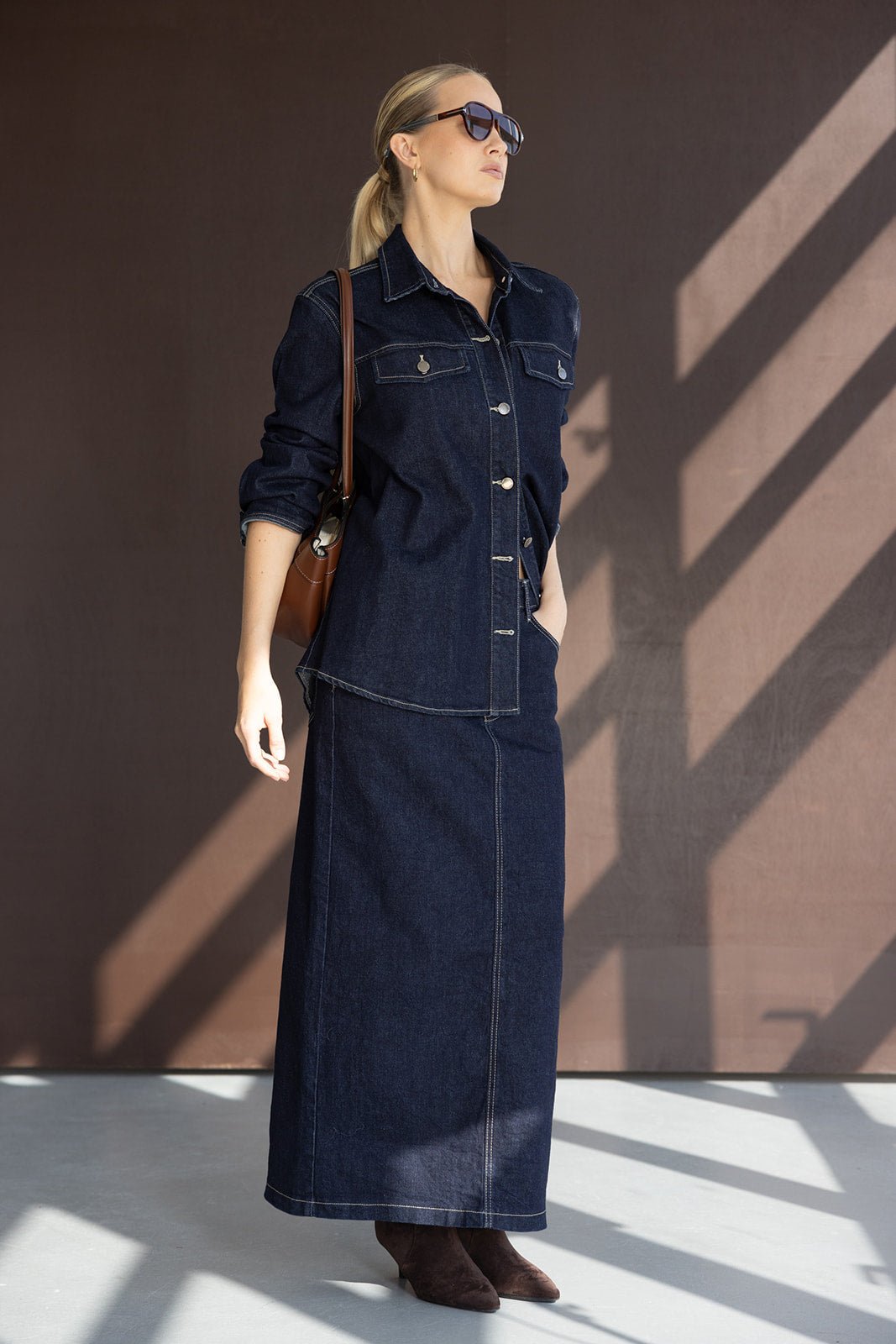 אישה לובשת חצאית ג'ינס ארוכה וז'קט ג'ינס כהה, עם משקפי שמש ותיק חום, אופנת נשים אלגנטית