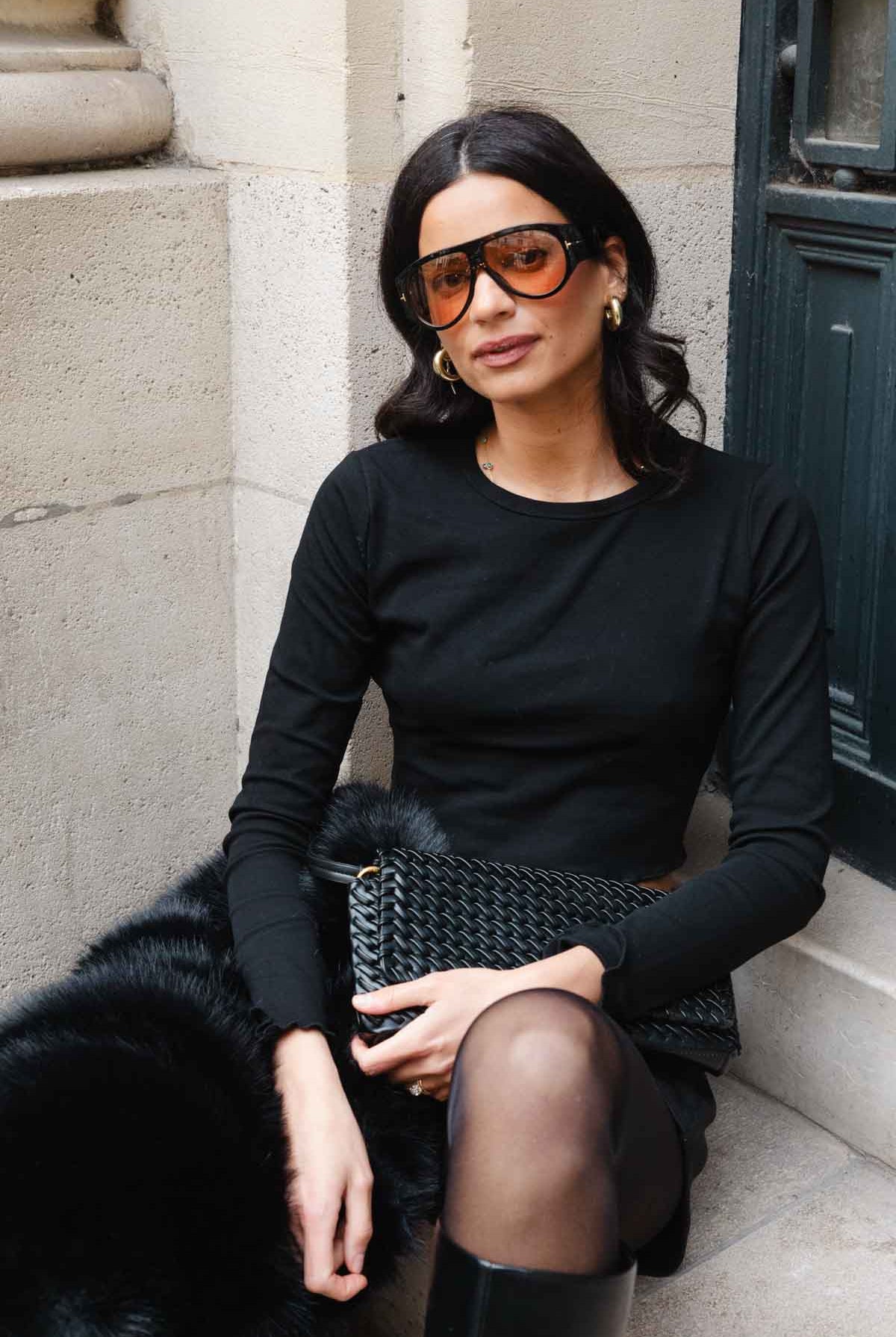 אישה עם משקפי שמש גדולים, לובשת חולצה שחורה וחצאית, מחזיקה תיק קלוע, יושבת ברחוב אופנתי