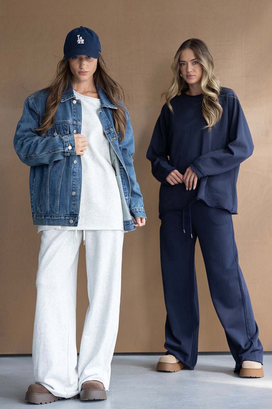 שתי נשים לובשות טרנינגים אופנתיים, אחת עם מעיל ג׳ינס וכובע, רקע חום מינימליסטי