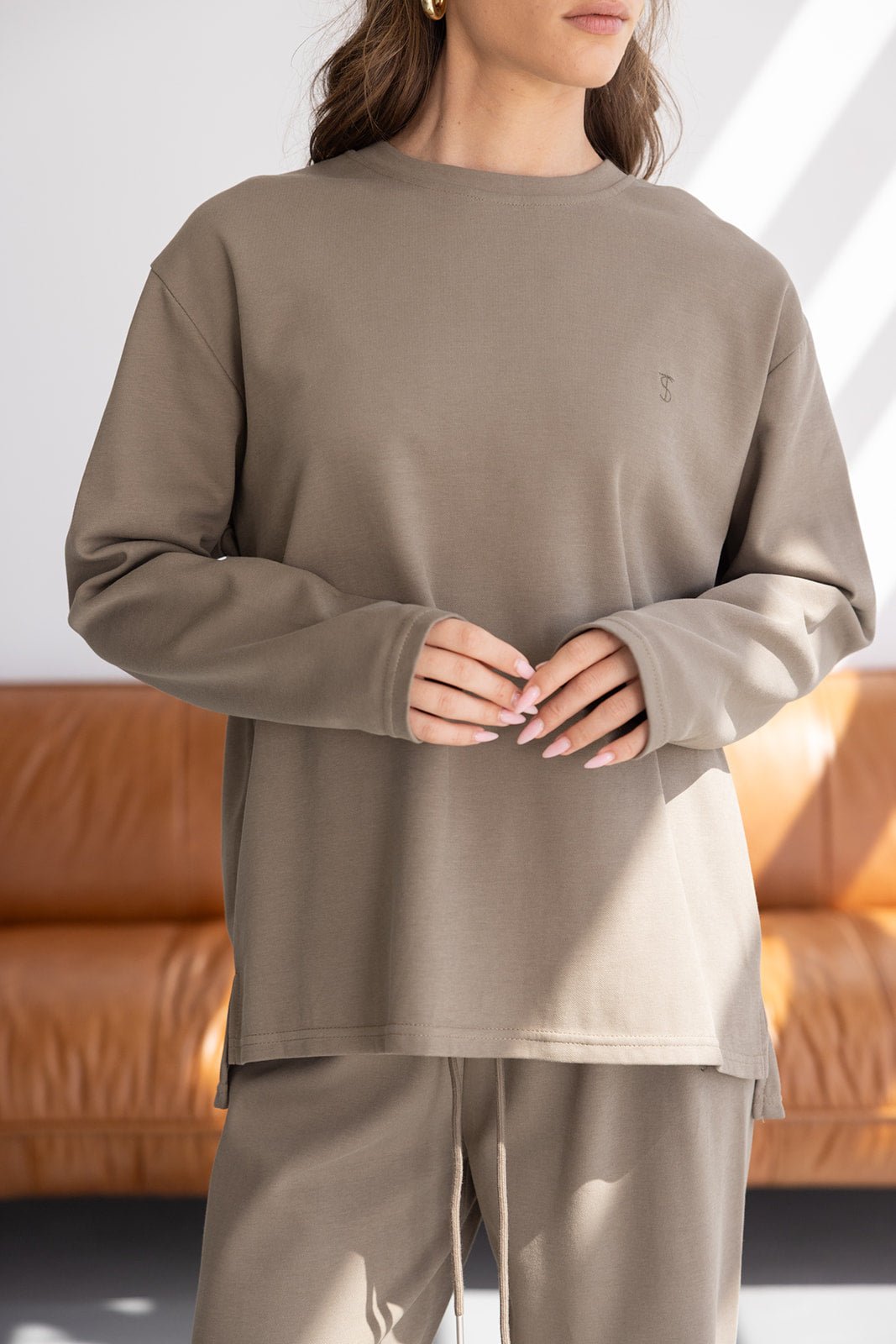 דוגמנית לובשת סט פוטר נשים בצבע בז' עם שרוולים ארוכים בגזרת אוברסייז, יושבת על ספה חומה