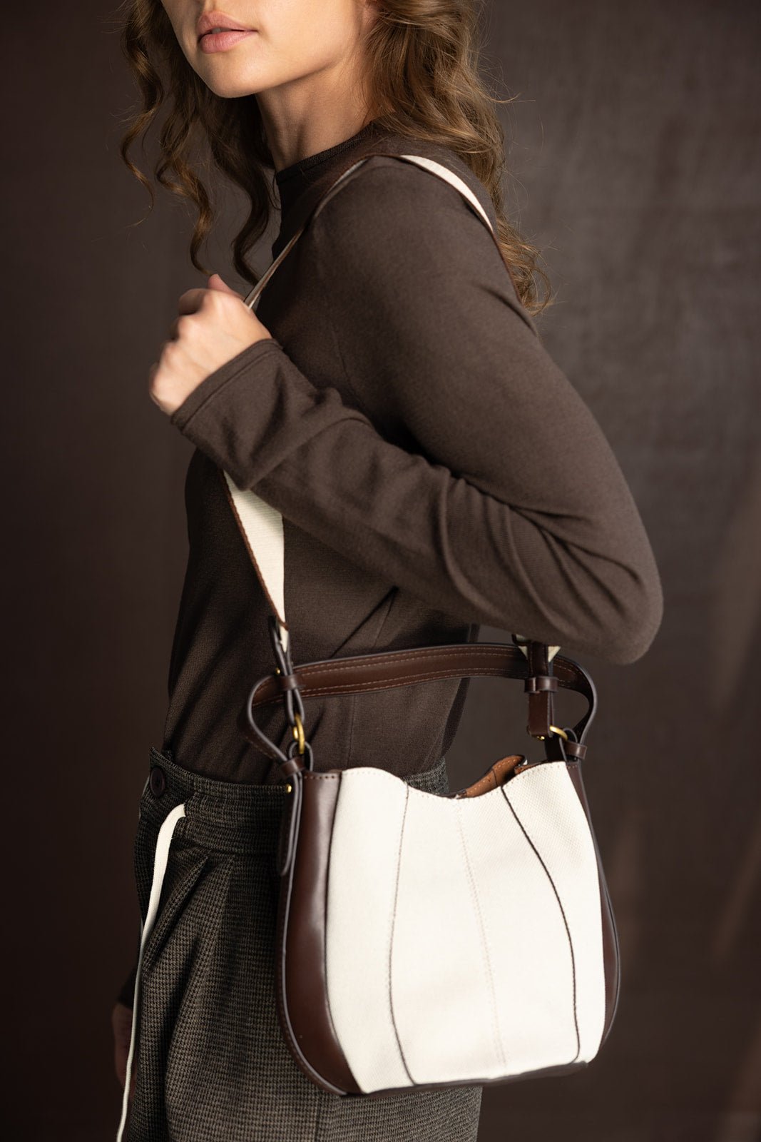 אישה עם חולצה חומה מחזיקה תיק צד אופנתי מבד לבן עם קצוות חומים, רקע כהה
