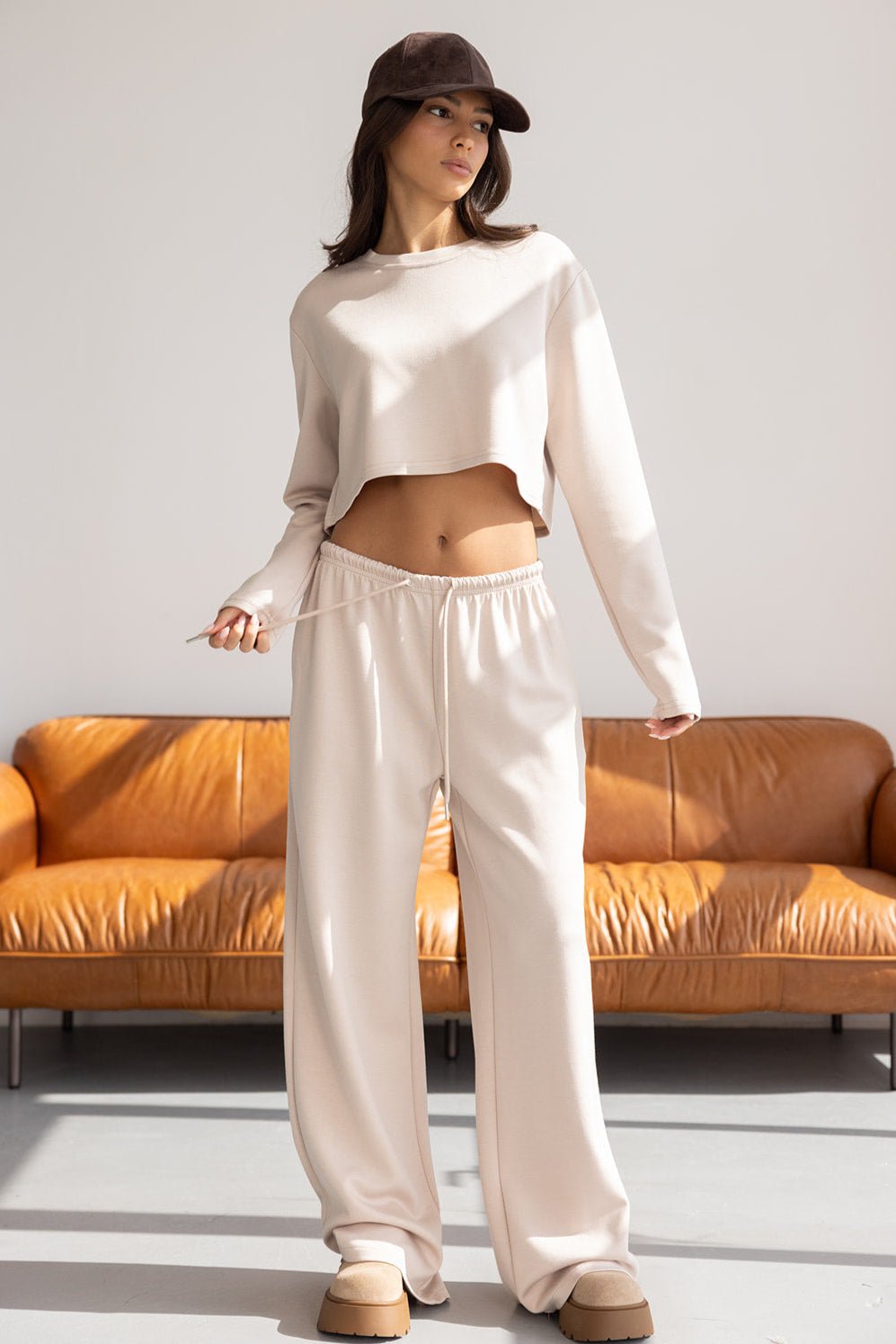 אישה עם חולצת קרופ ארוכה ומכנסי טרנינג בז', כובע קלאסי, יושבת על ספה בסלון מואר