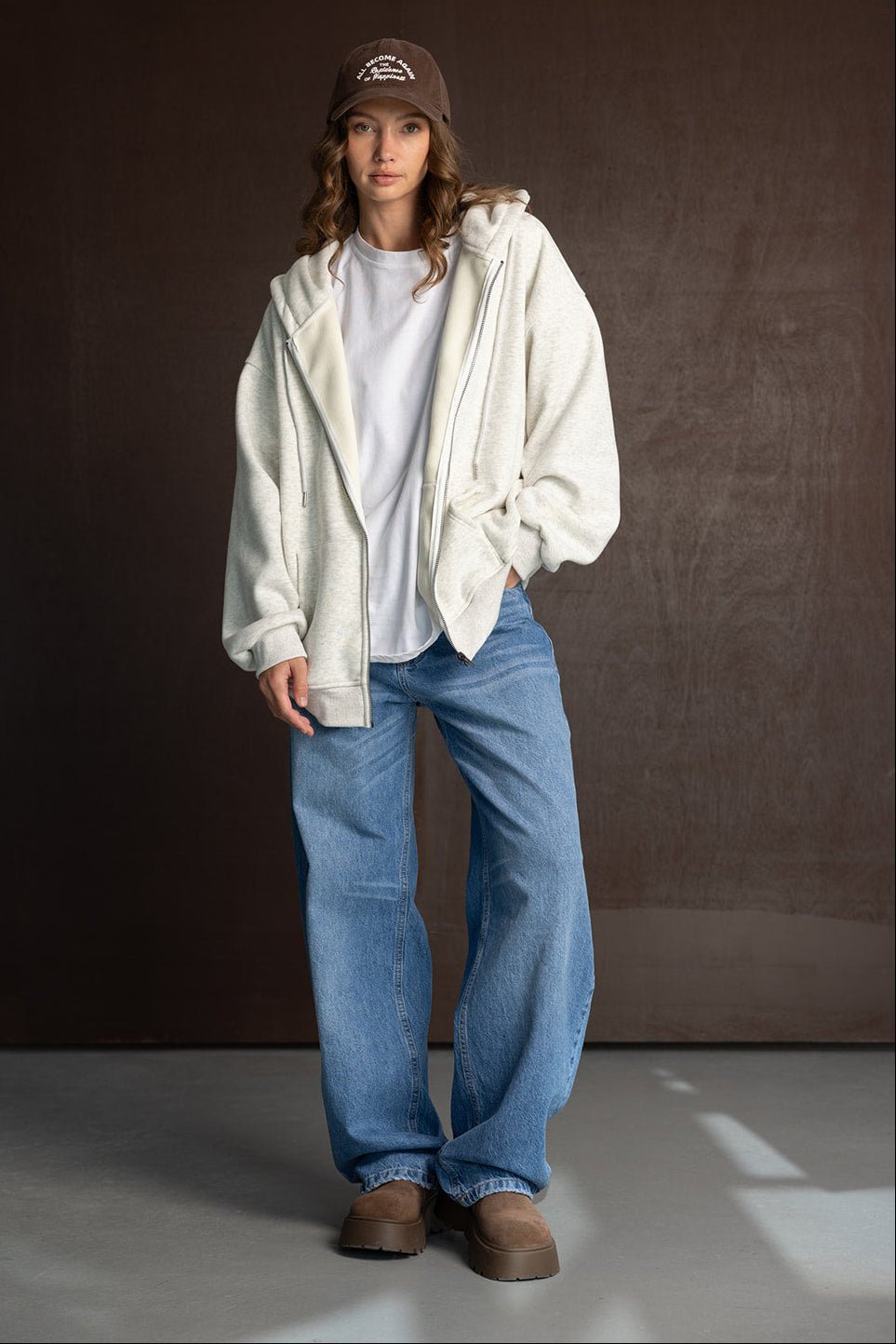 אישה לובשת קפוצ'ון אפור, ג'ינס כחול רחב וכובע חום, לוק קז'ואל אופנתי לחורף