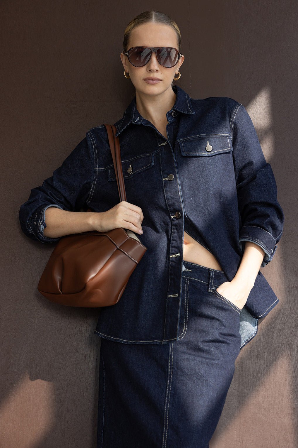 אישה עם חולצת ג'ינס כהה, חצאית ג'ינס, תיק חום ומשקפי שמש אופנתיים