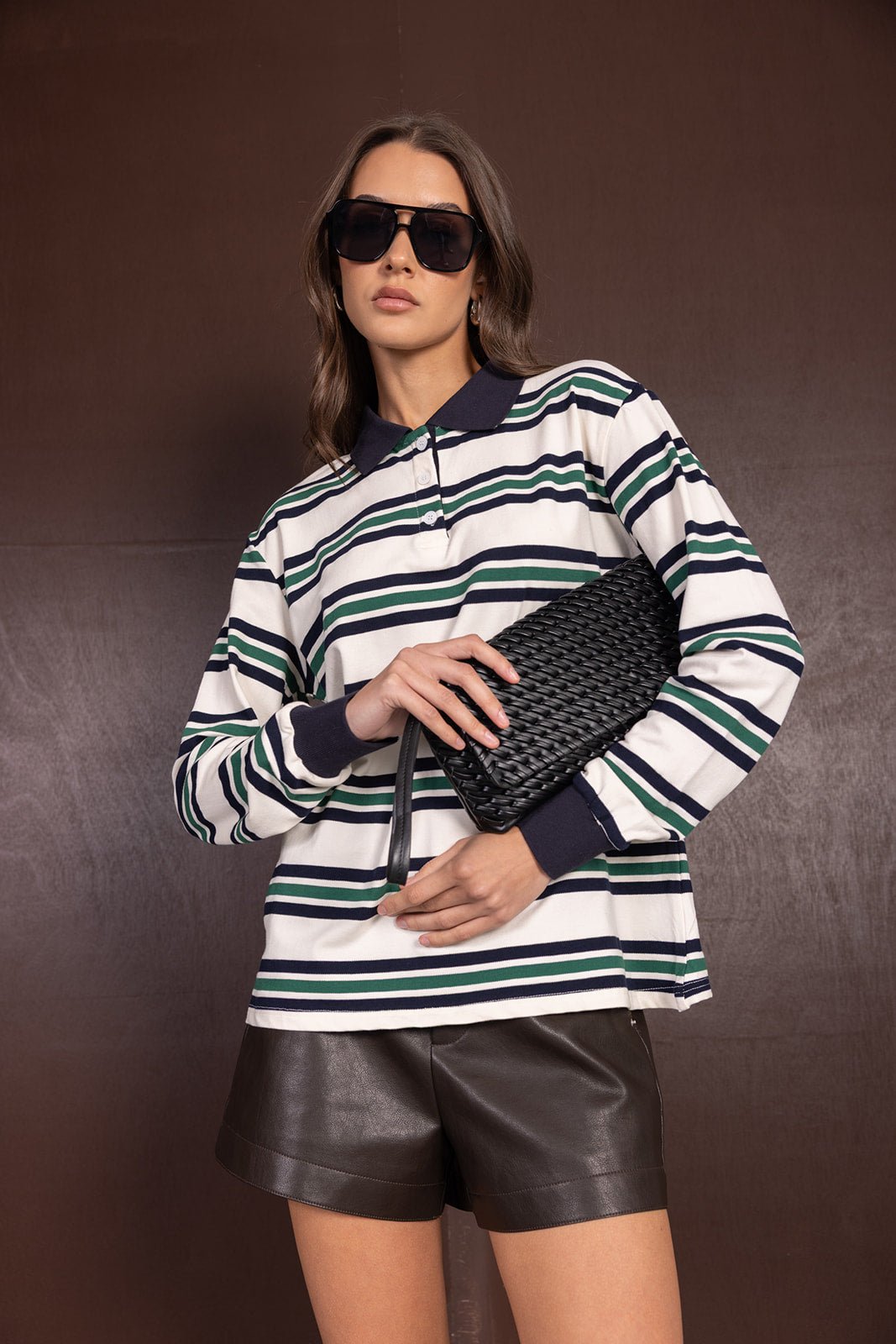 אישה עם חולצת פסים ארוכה, שורט עור שחור, תיק קלאץ' קלוע ומשקפי שמש אופנתיים