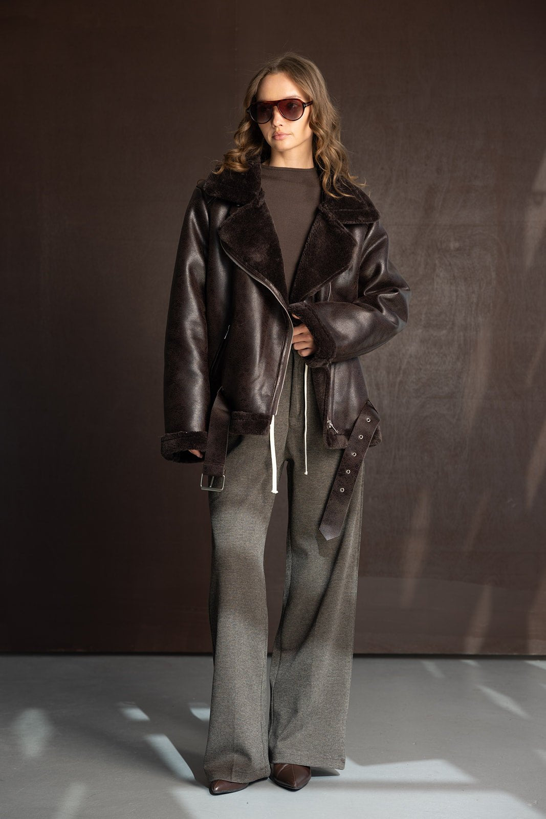 דוגמנית עם מעיל עור חום, משקפי שמש חומים ומכנסיים רחבים אופנתיים, רקע כהה