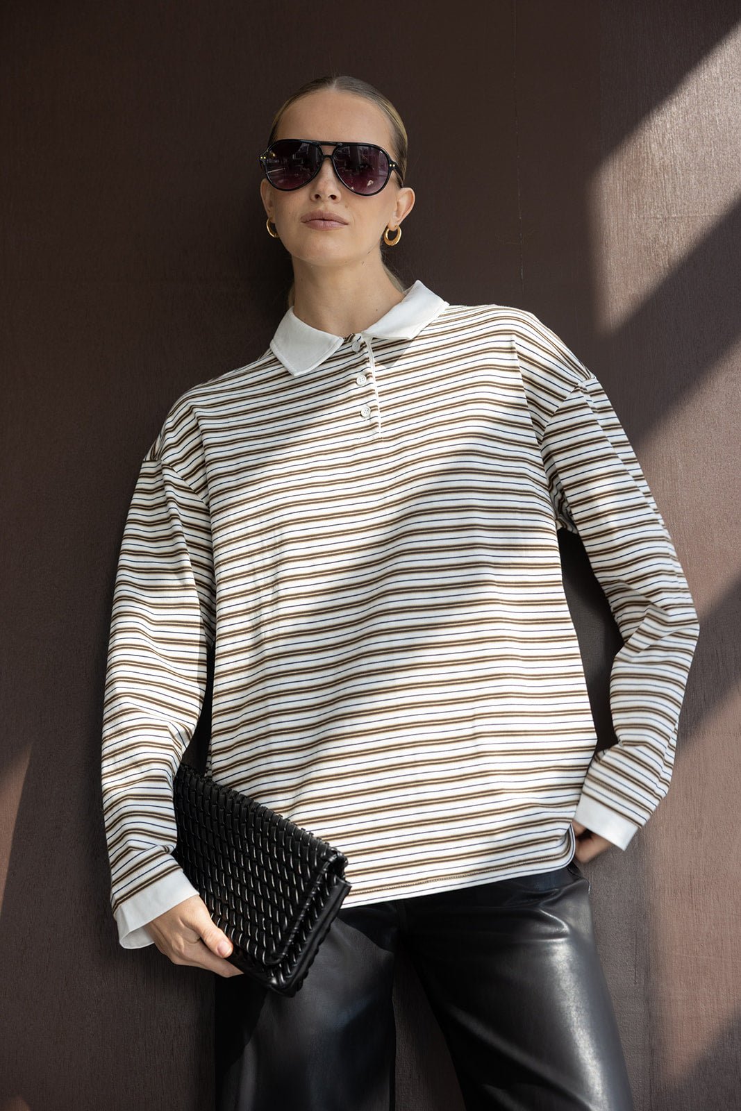 אישה עם חולצת פסים ארוכה, מכנסי עור שחורים, משקפי שמש ותיק קלאץ' שחור – אופנת נשים טרנדית