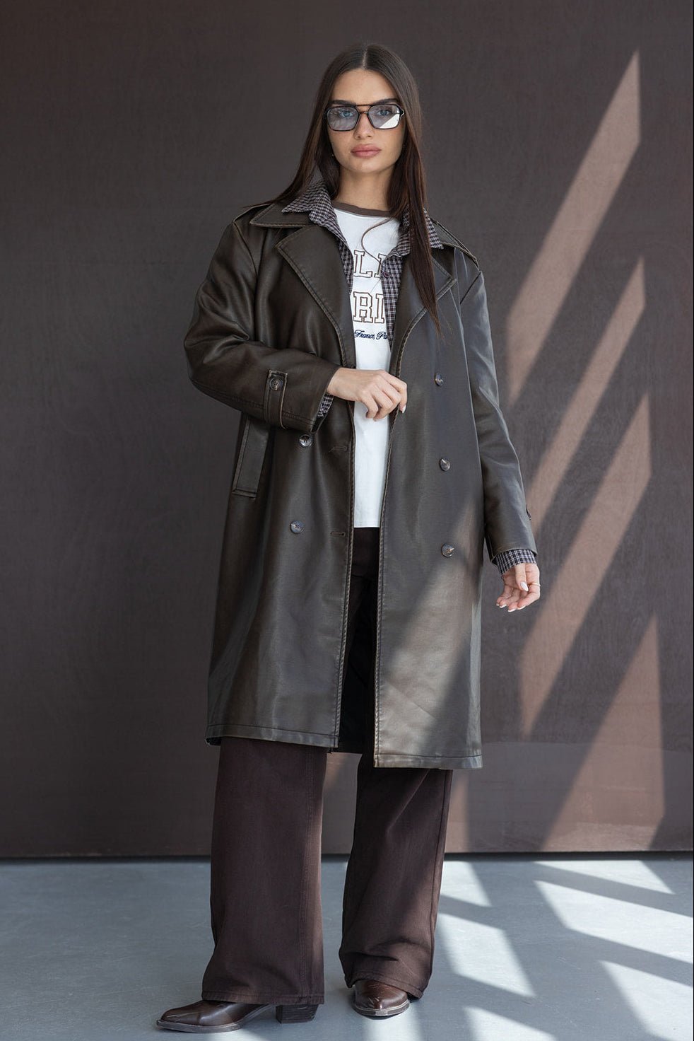 אשה עם מעיל עור חום, משקפי שמש, חולצת טריקו ומכנסיים רחבים באופנת חורף לנשים