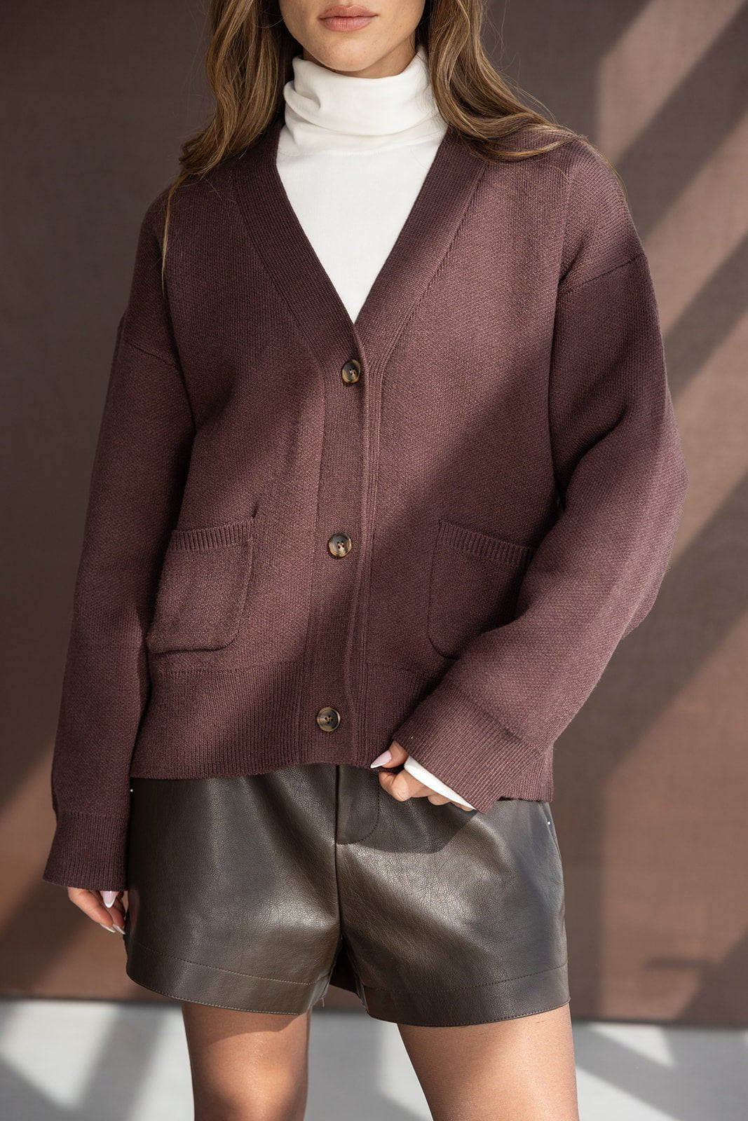 אישה לובשת קרדיגן סגול כהה עם כפתורים, גולף לבן ומכנסי עור חומים, אופנת נשים חורף