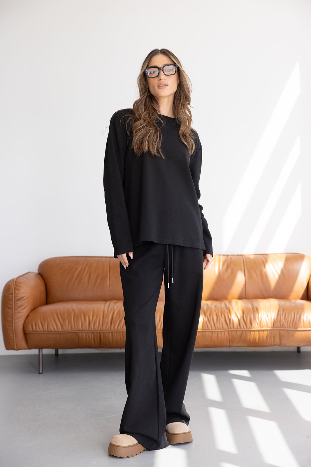 אישה מדגמנת סט טרנינג שחור אופנתי, משקפי שמש, ספה חומה ברקע, אופנת נשים טרנדית