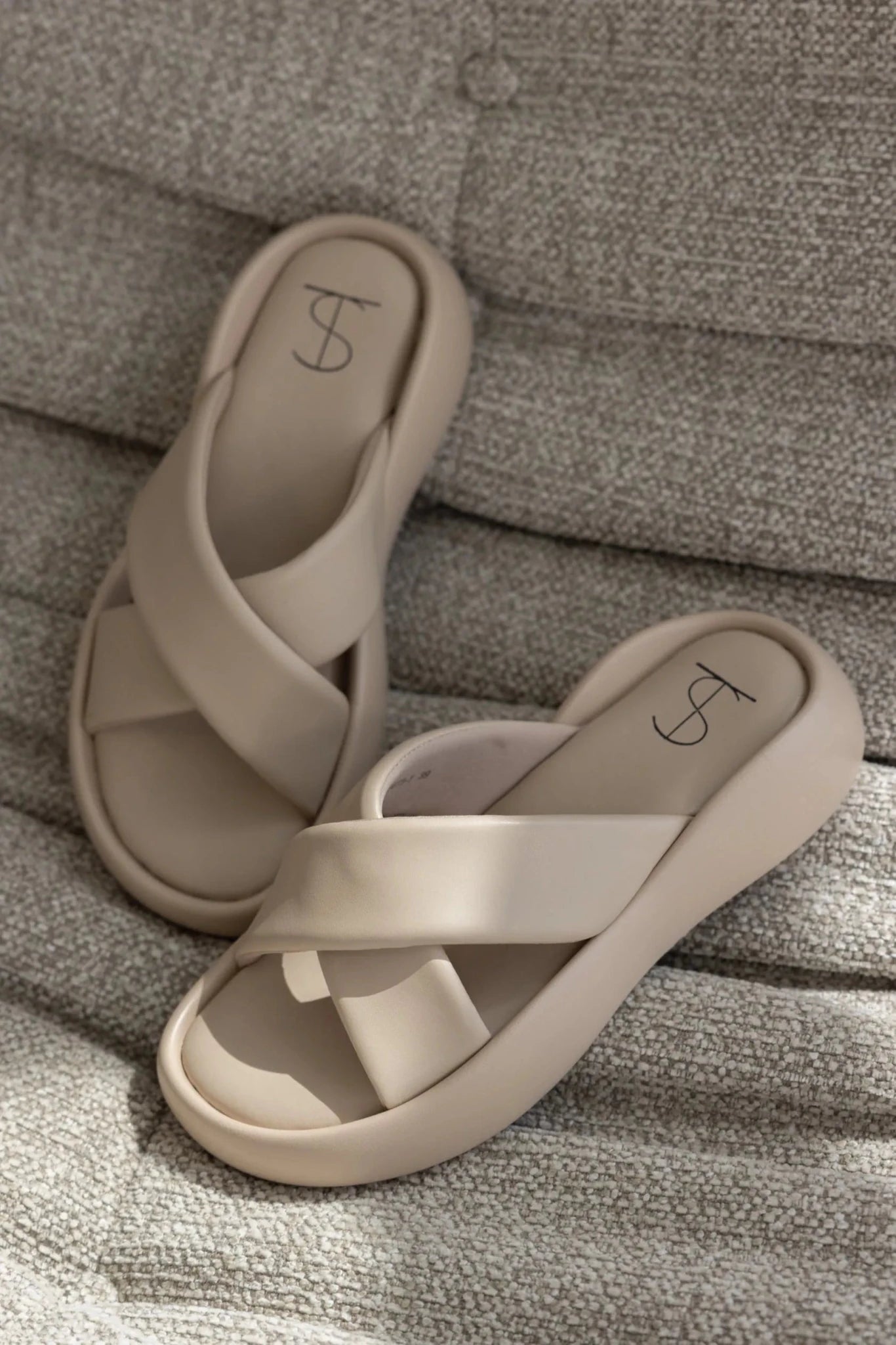 כפכפי נשים בצבע בז' עם רצועות שלובות, מונחים על ספה מבד בהיר, אופנת קיץ נוחה