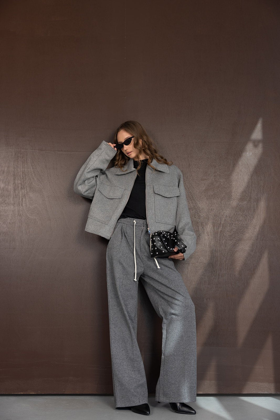 אישה לובשת חליפת מכנסיים מחויטת אפורה בסגנון קז'ואל, חולצה שחורה, משקפי שמש ותיק שחור, מראה אופנתי לנשים צעירות.