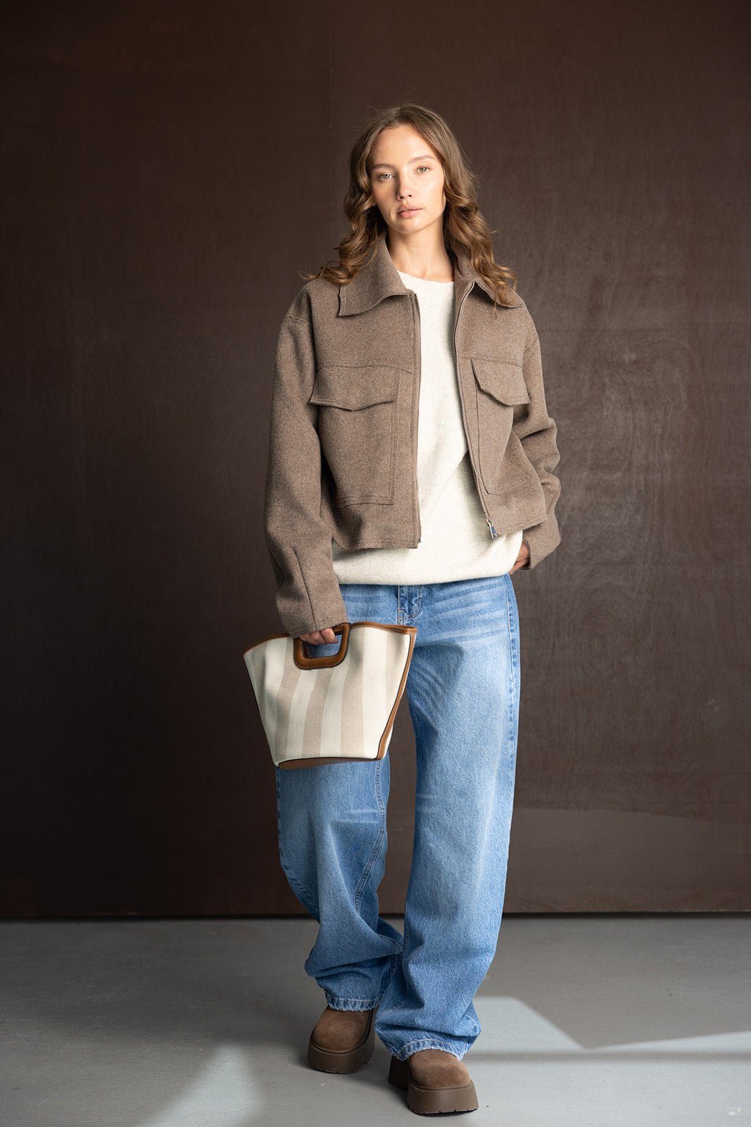 אישה עם ז'קט חום, ג'ינס רחב, סוודר לבן, תיק בד ונעליים חומות – אופנת חורף לנשים