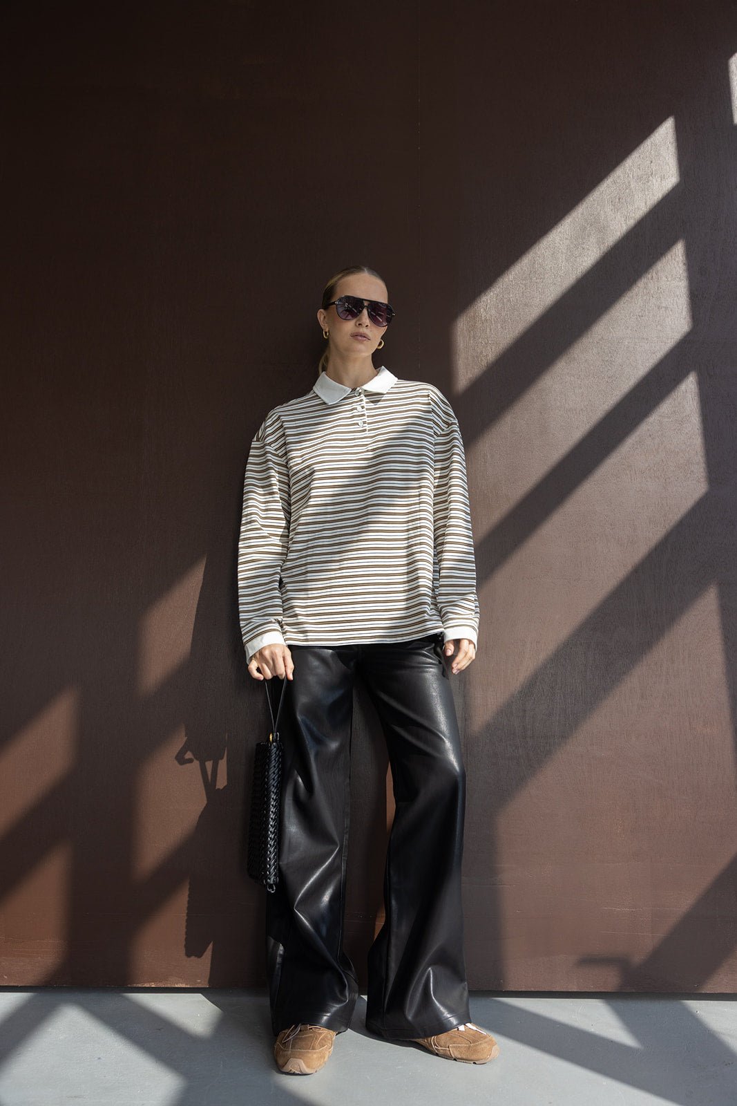 אישה עם חולצת פסים ארוכה ומכנסי דמוי עור שחורים, משקפי שמש ואביזרי אופנה, מראה אופנתי צעיר