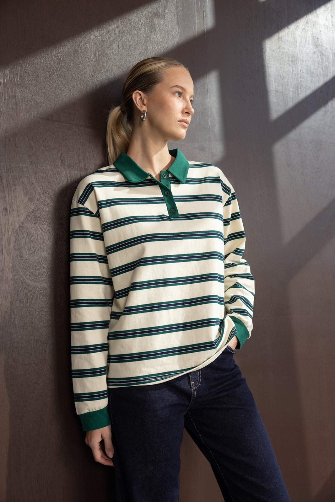 אישה צעירה לבושה חולצת פסים ארוכה עם צווארון ירוק ומכנסי ג'ינס, עומדת לצד קיר