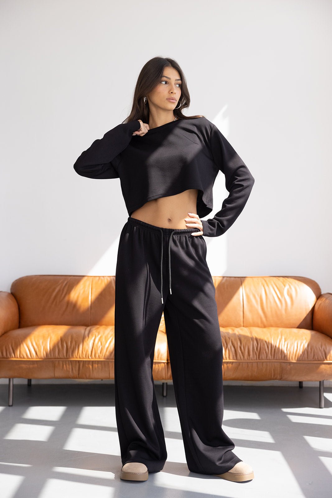 אישה לובשת סט טרנינג שחור אופנתי עם סווטשירט קצוץ ומכנסיים רחבים, עומדת בסלון מואר