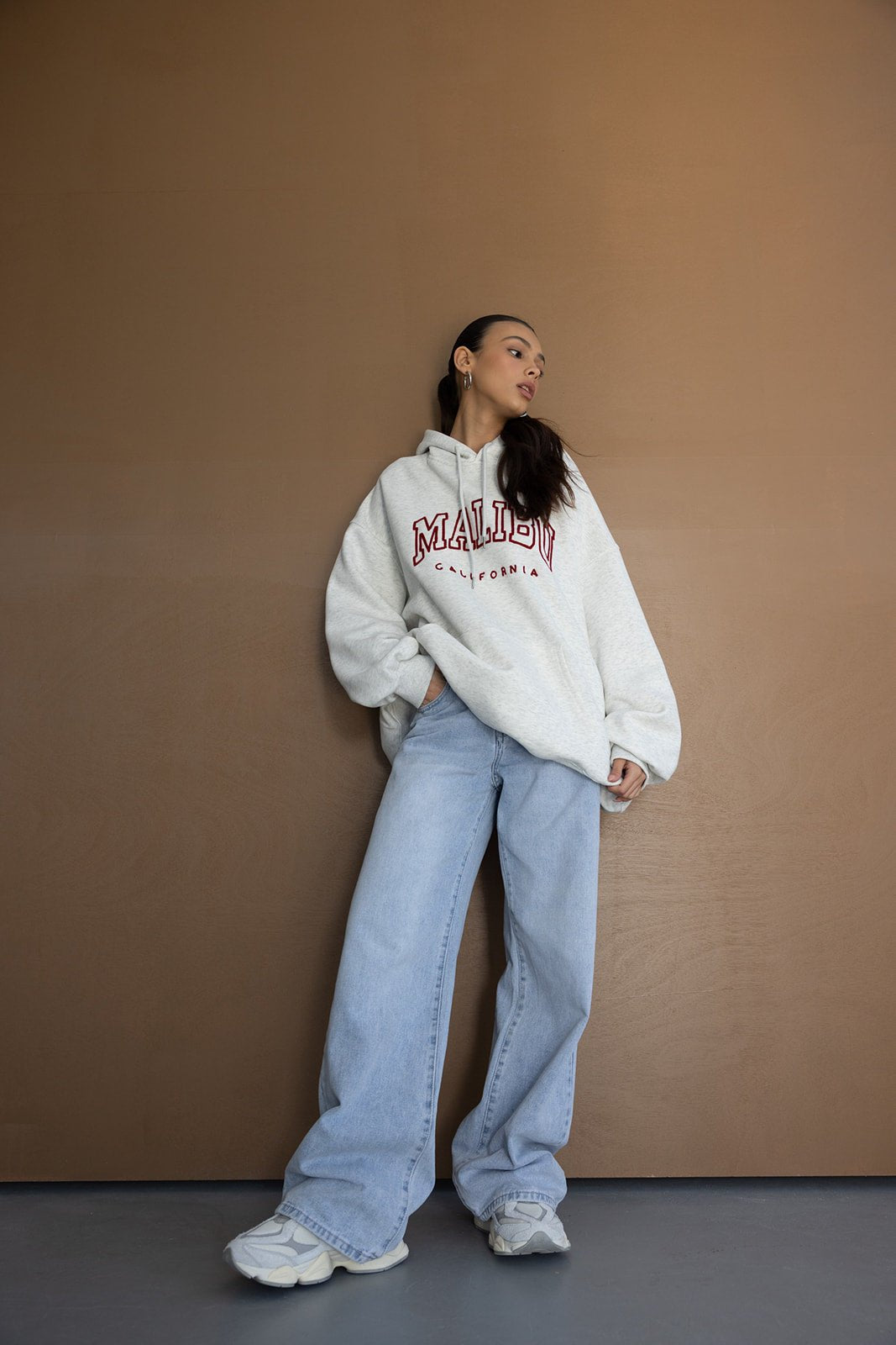 אישה צעירה לובשת סווטשירט אפור עם כיתוב 'Malibu', ג'ינס רחב ונעלי סניקרס טרנדיים, לוק אופנתי לנערות