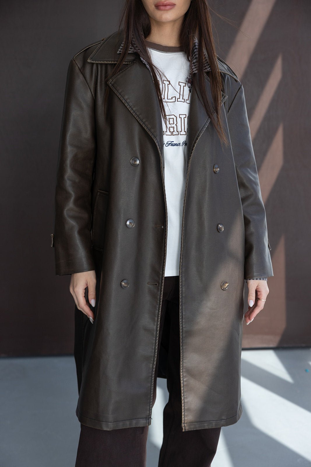 אישה עם מעיל טרנץ' ארוך דמוי עור בצבע חום כהה, חולצה לבנה עם הדפס, סטייל חורפי אופנתי