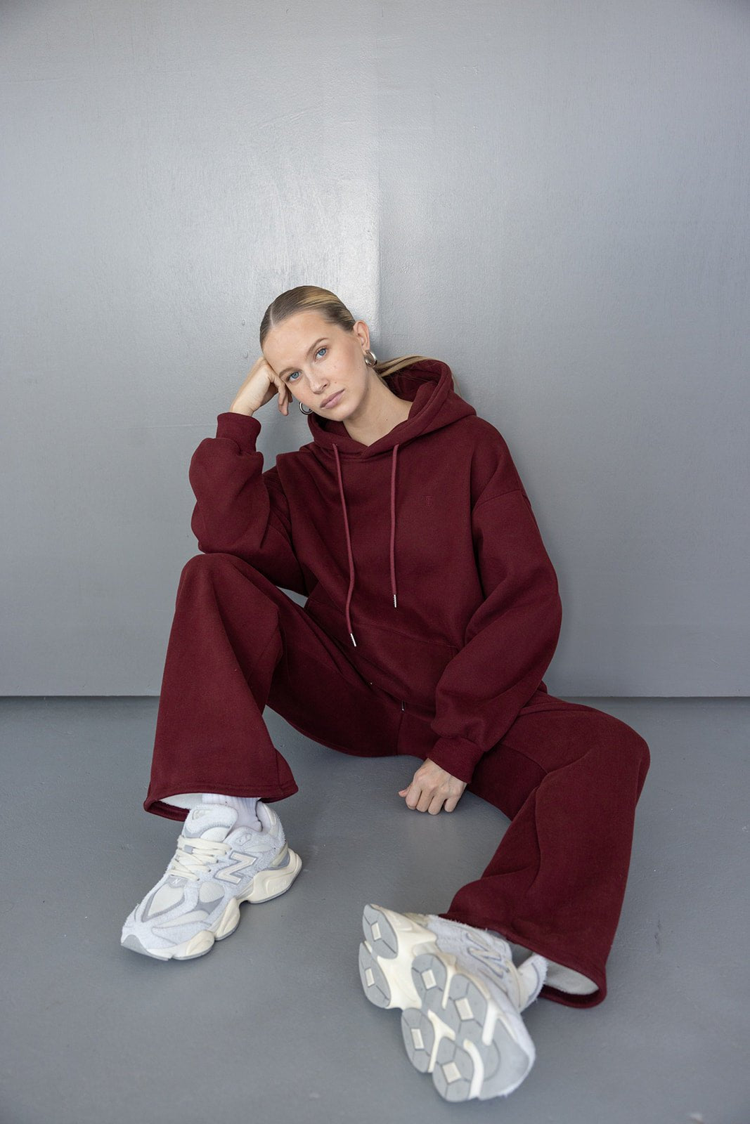 דוגמנית יושבת על הרצפה בחליפת טרנינג בורדו אופנתית ונעלי סניקרס לבנות, לוק ספורטיבי לנשים