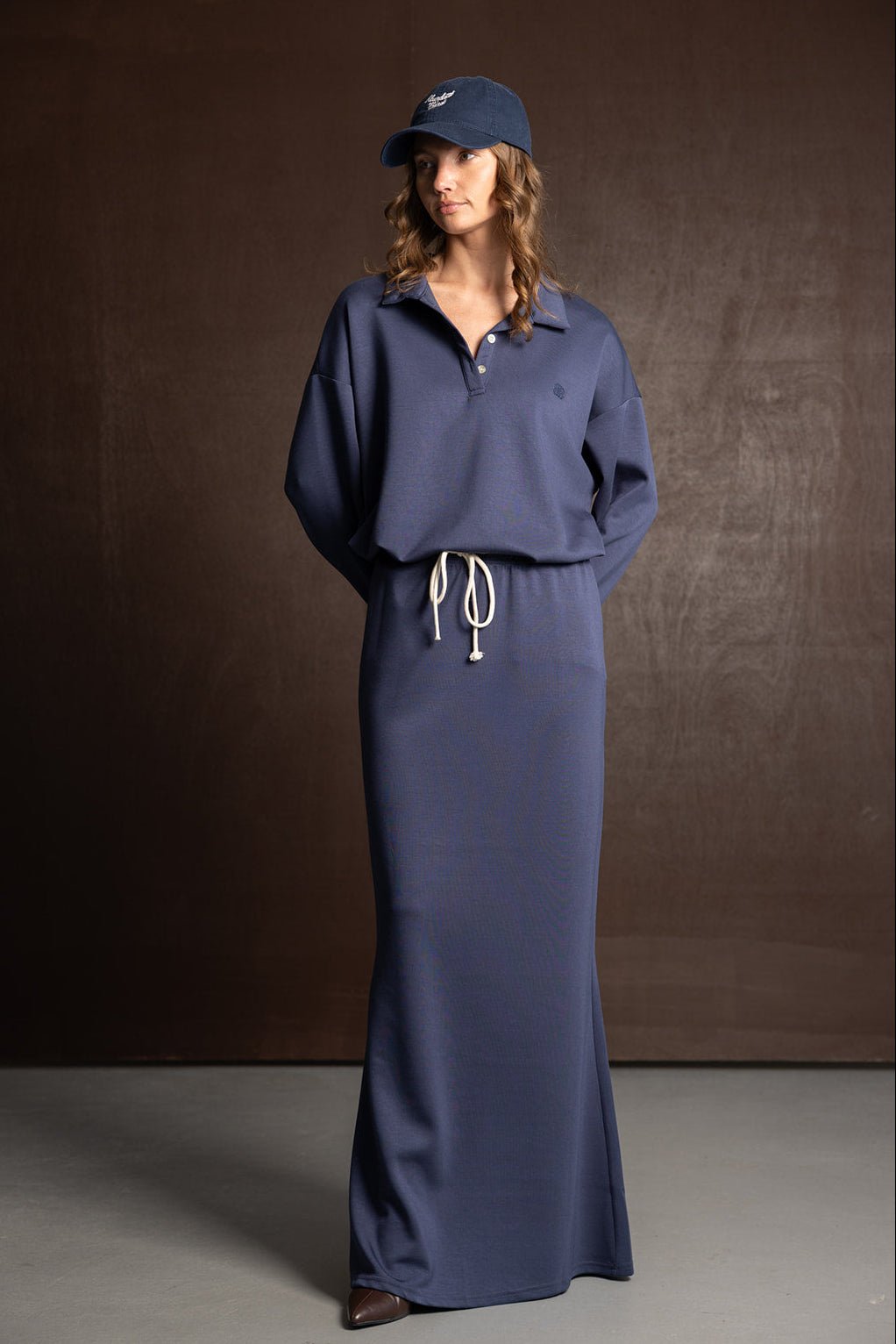 אישה עם שמלה כחולה ארוכה, שרוולים ארוכים, כובע מצחייה כחול, מראה אופנה צעיר ועדכני