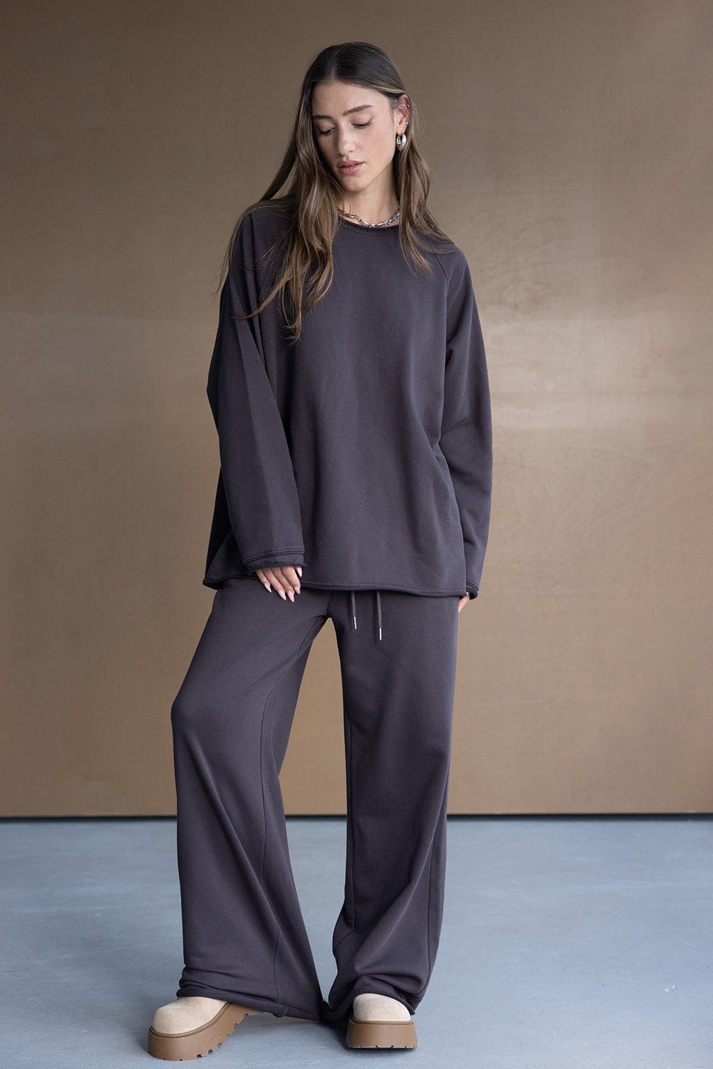אישה לובשת סט טרנינג אופנתי בצבע אפור כהה, חולצה ארוכה ומכנסיים רחבים, עם נעליים פלטרפורמה, רקע ניטרלי