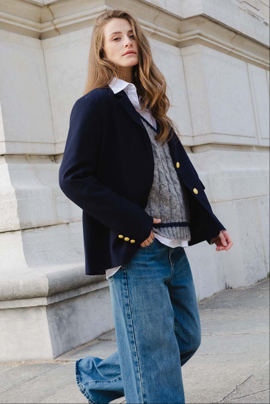 אישה צעירה לובשת ז'קט כחול כהה עם כפתורים זהב, סוודר אפור ומכנסי ג'ינס רחבים, אופנת רחוב לנשים